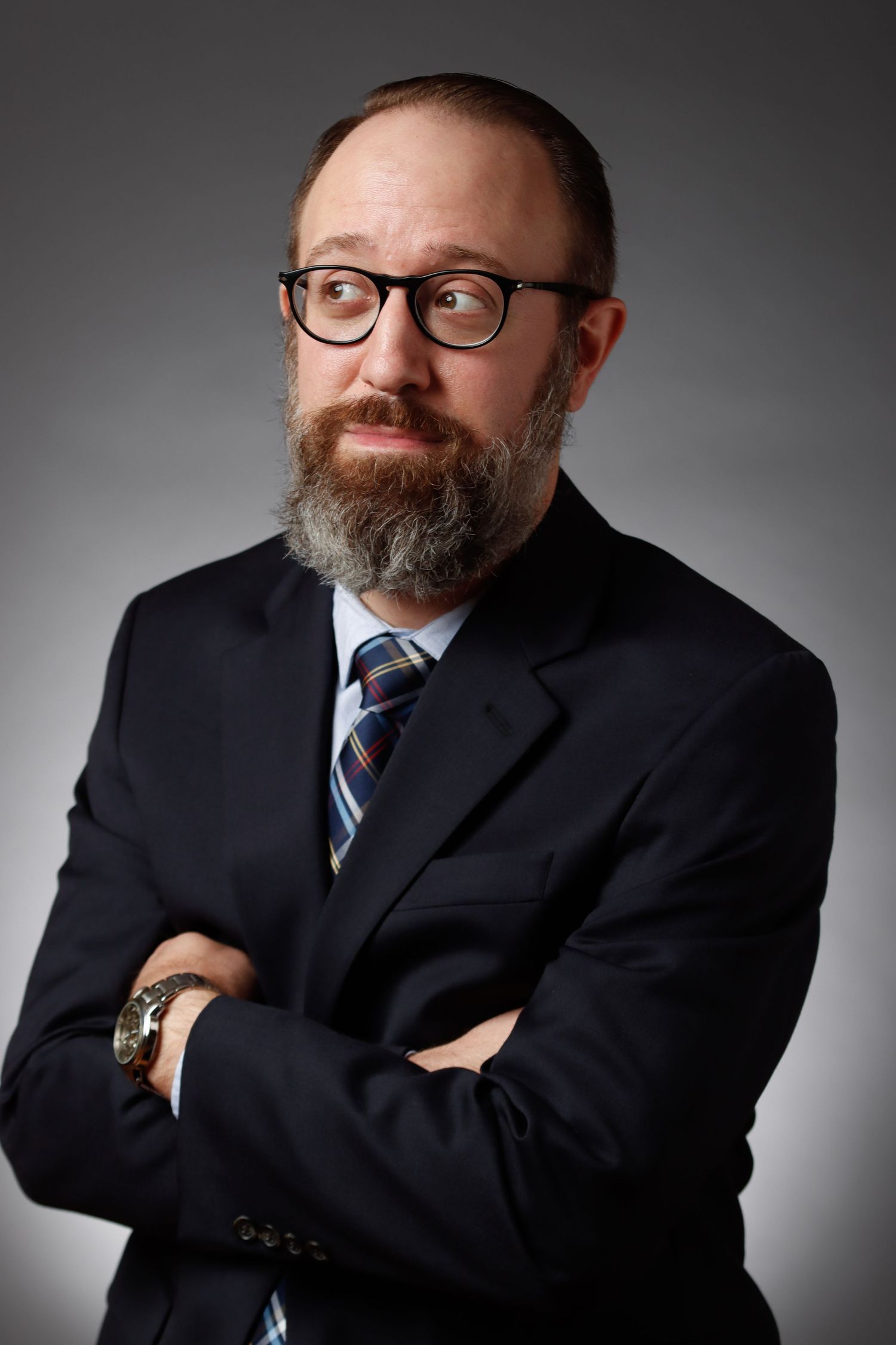 Bearded man in dark suit with glasses posing with arms crossed in studio.