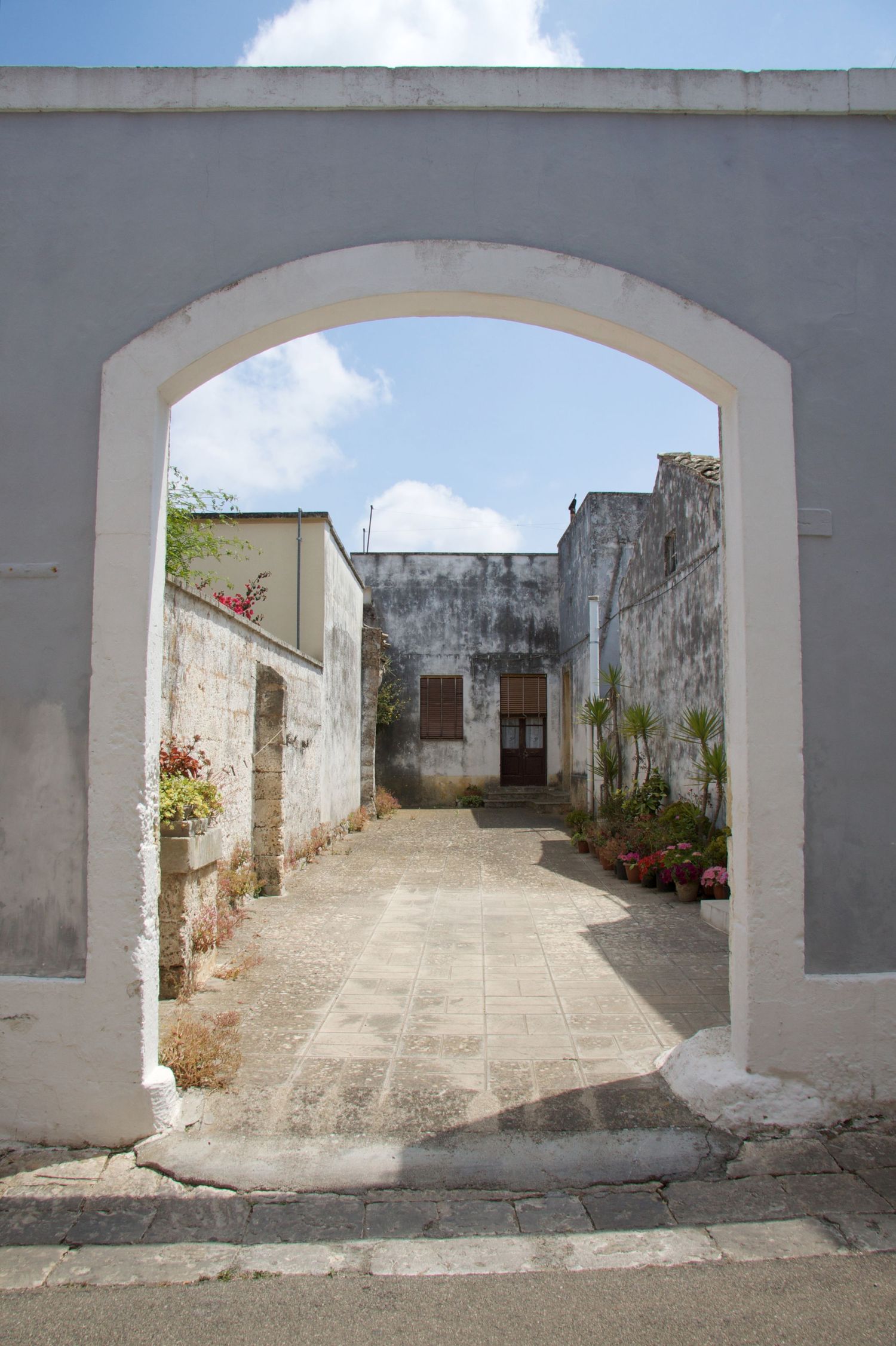 White stucco archway entrance leading to a sunlit Mediterranean courtyard with flowering plants and stone walls.