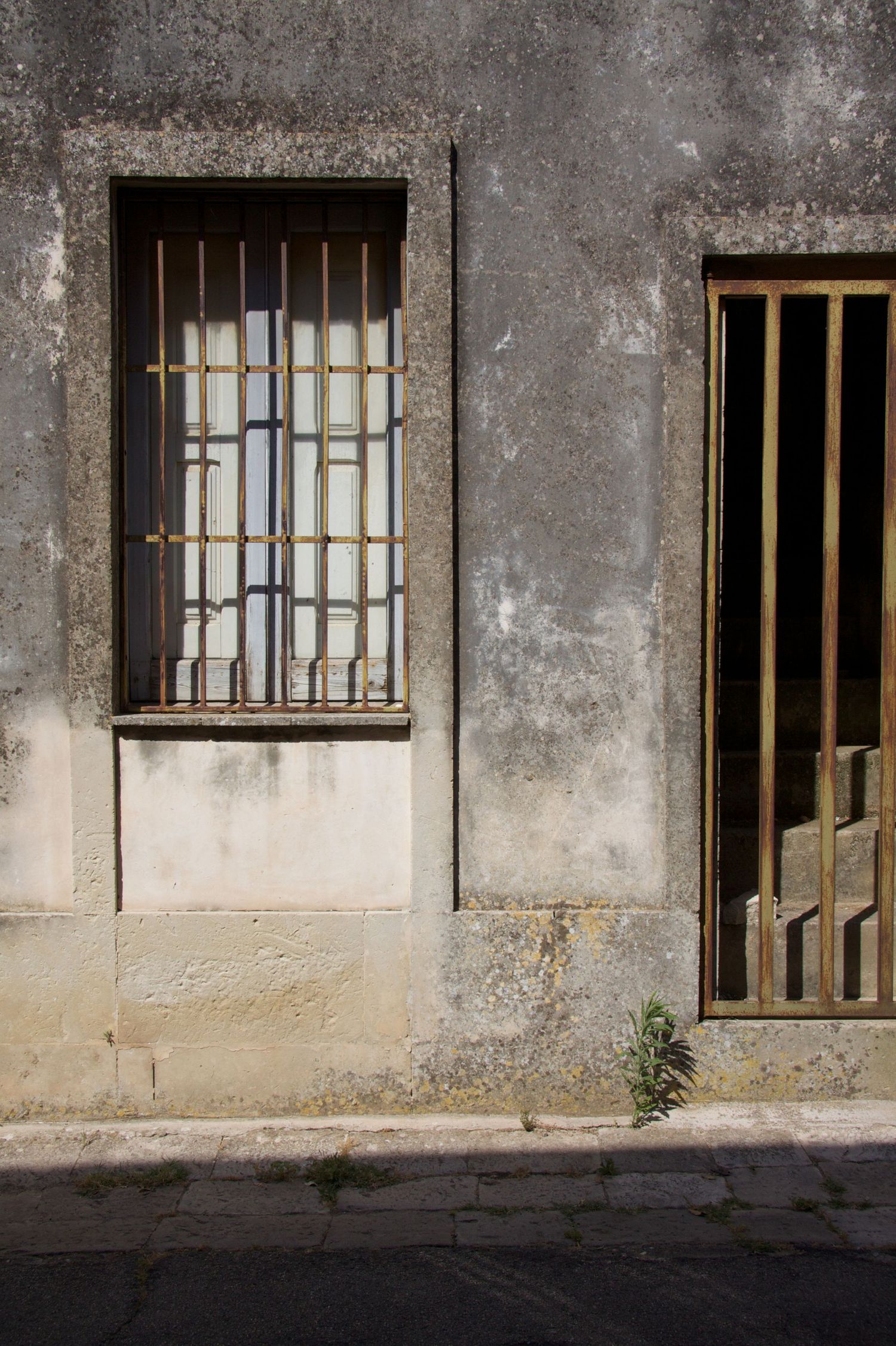 Weathered gray concrete building facade with rusty iron bar window grate and door on a sunny urban street.