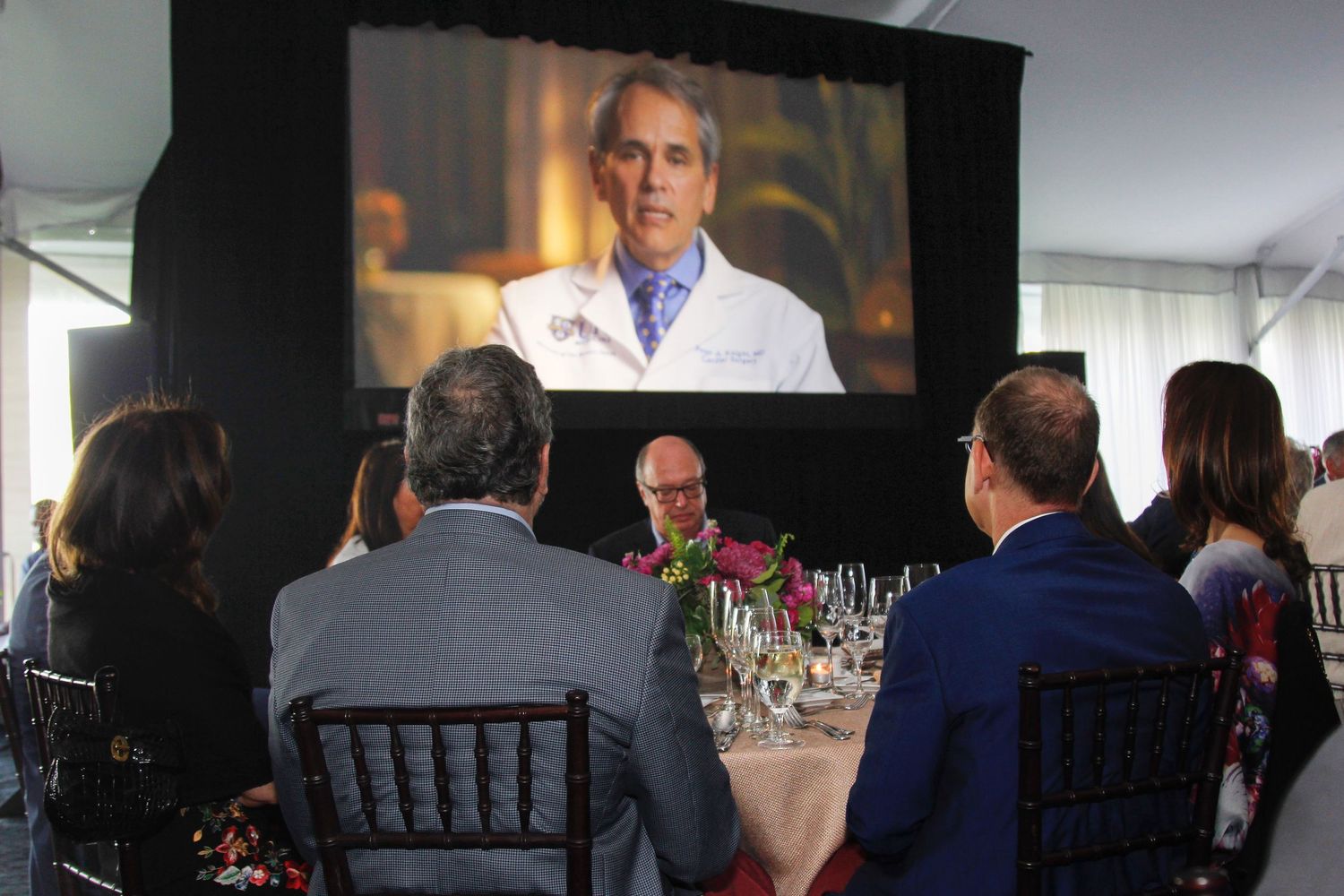 Guests watch a doctor speaking on a large screen during an intimate formal dinner presentation.