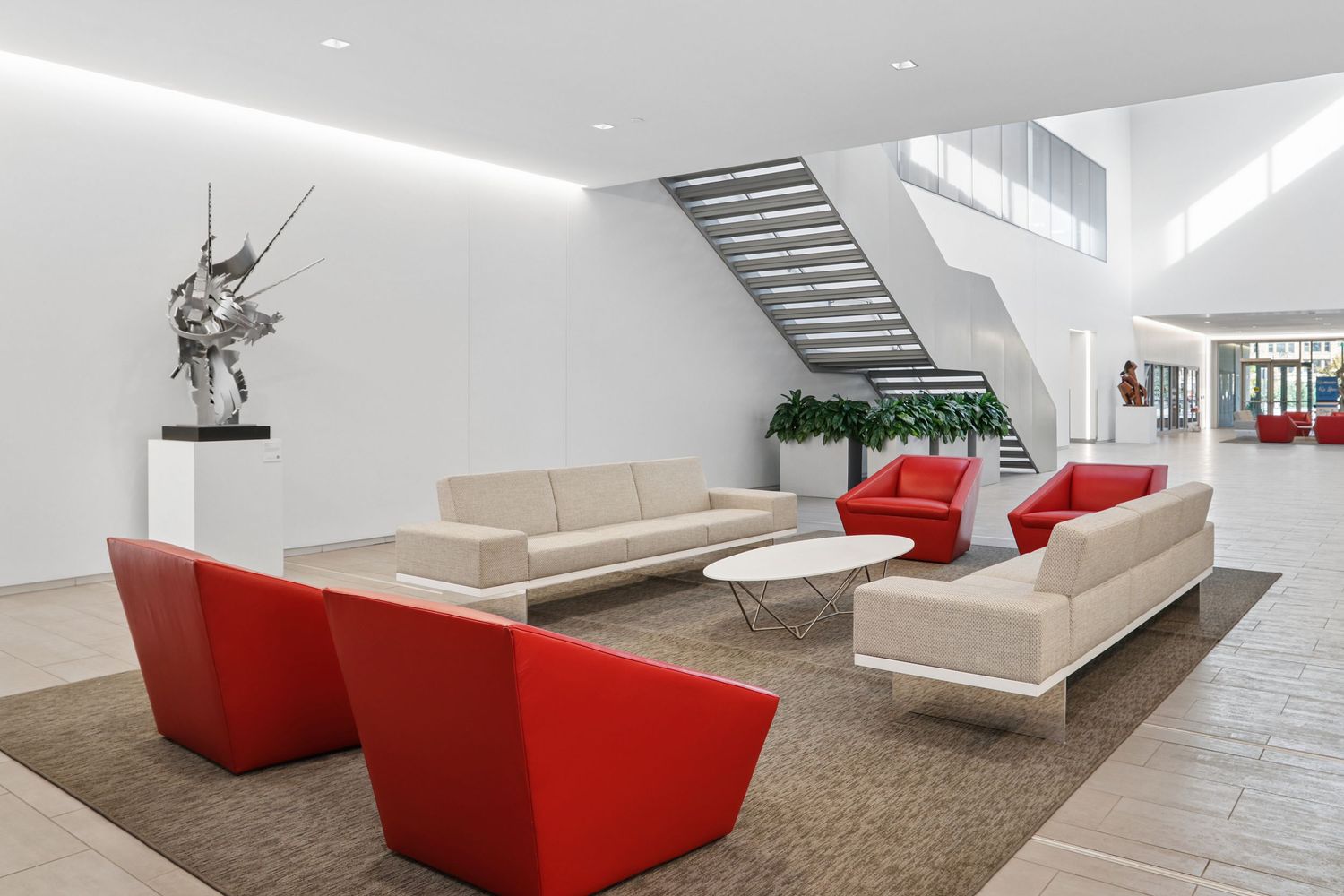 Modern corporate lobby with red accent chairs, beige sofas, white walls, floating staircase, and contemporary sculpture on display.