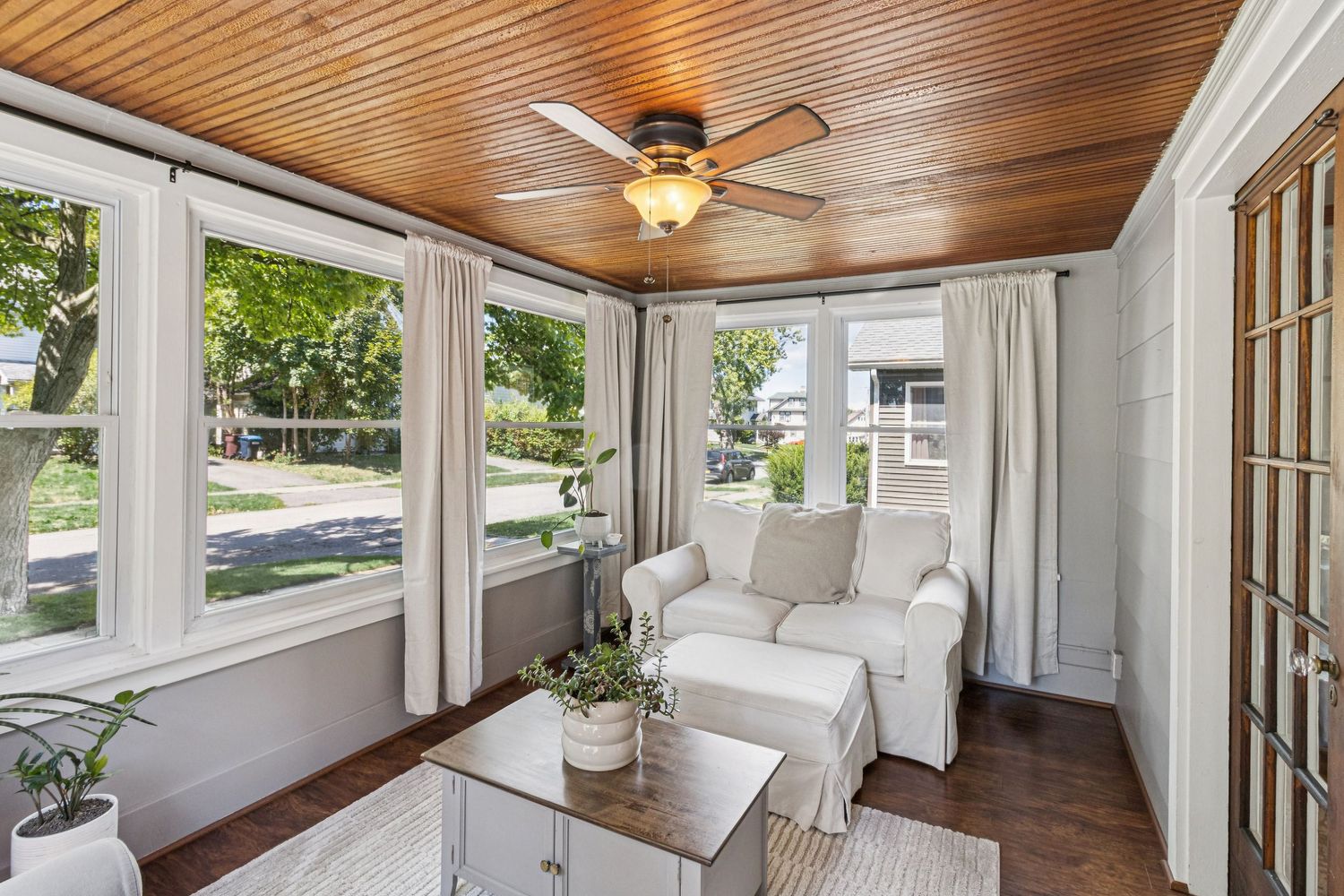 Bright sunroom with wood ceiling, ceiling fan, white furniture, large windows, and hardwood floors.