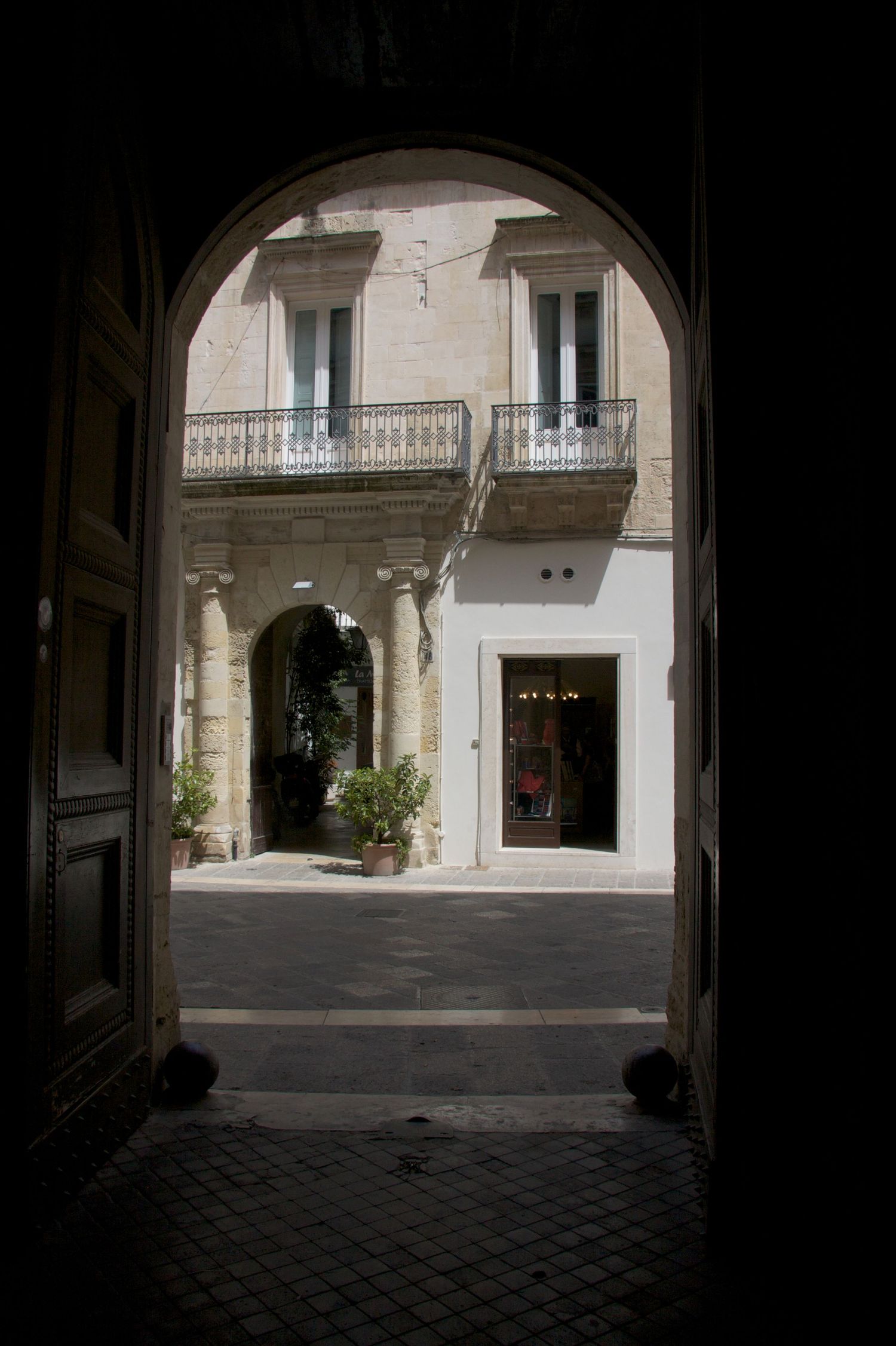 Dark archway entrance framing a sunlit Italian courtyard with balcony and arched colonnade.