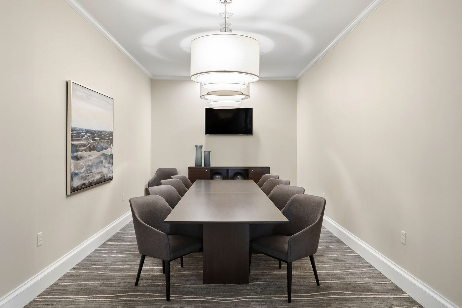 Modern conference room with a long dark wood table, gray chairs, pendant light, wall-mounted TV, and neutral beige walls.
