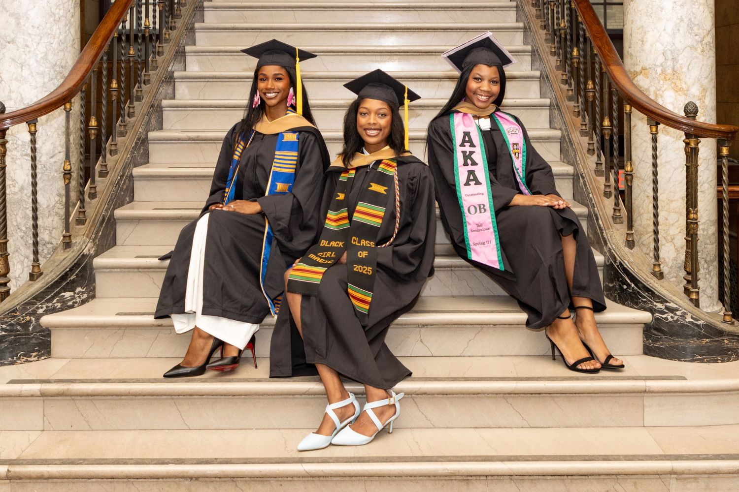 Three graduates in caps and gowns sitting on marble staircase at graduation.