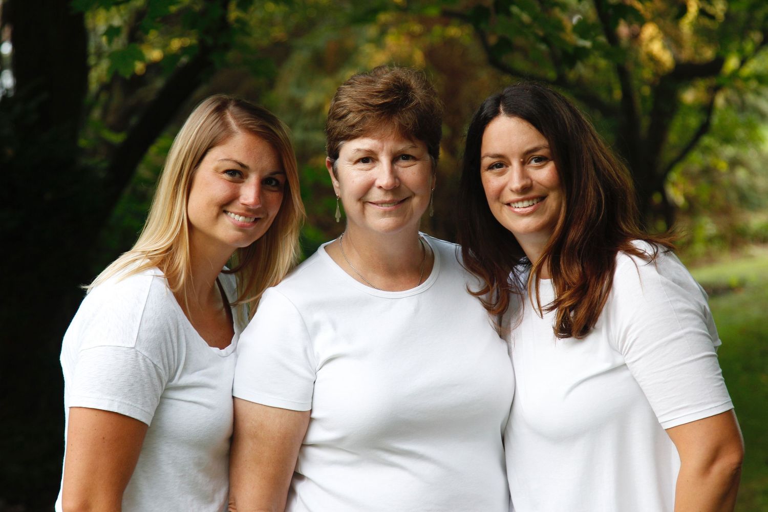Three women in matching white shirts smiling together in an outdoor green setting.