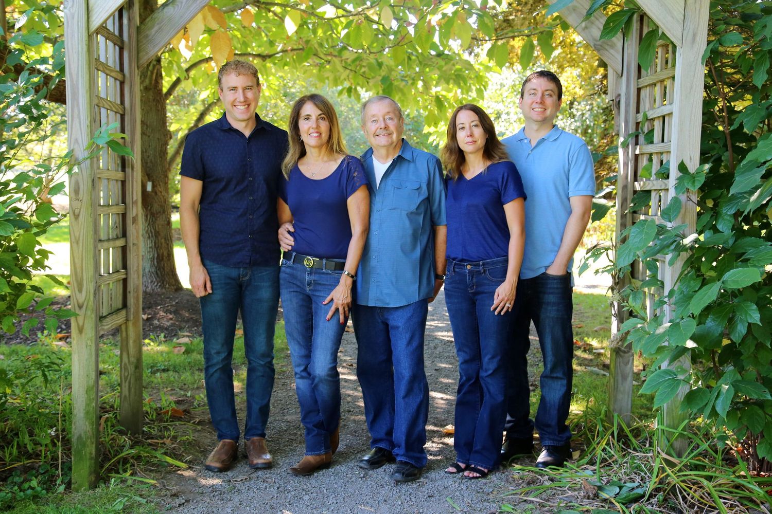 Family of five smiling together under garden arbor surrounded by lush green foliage.