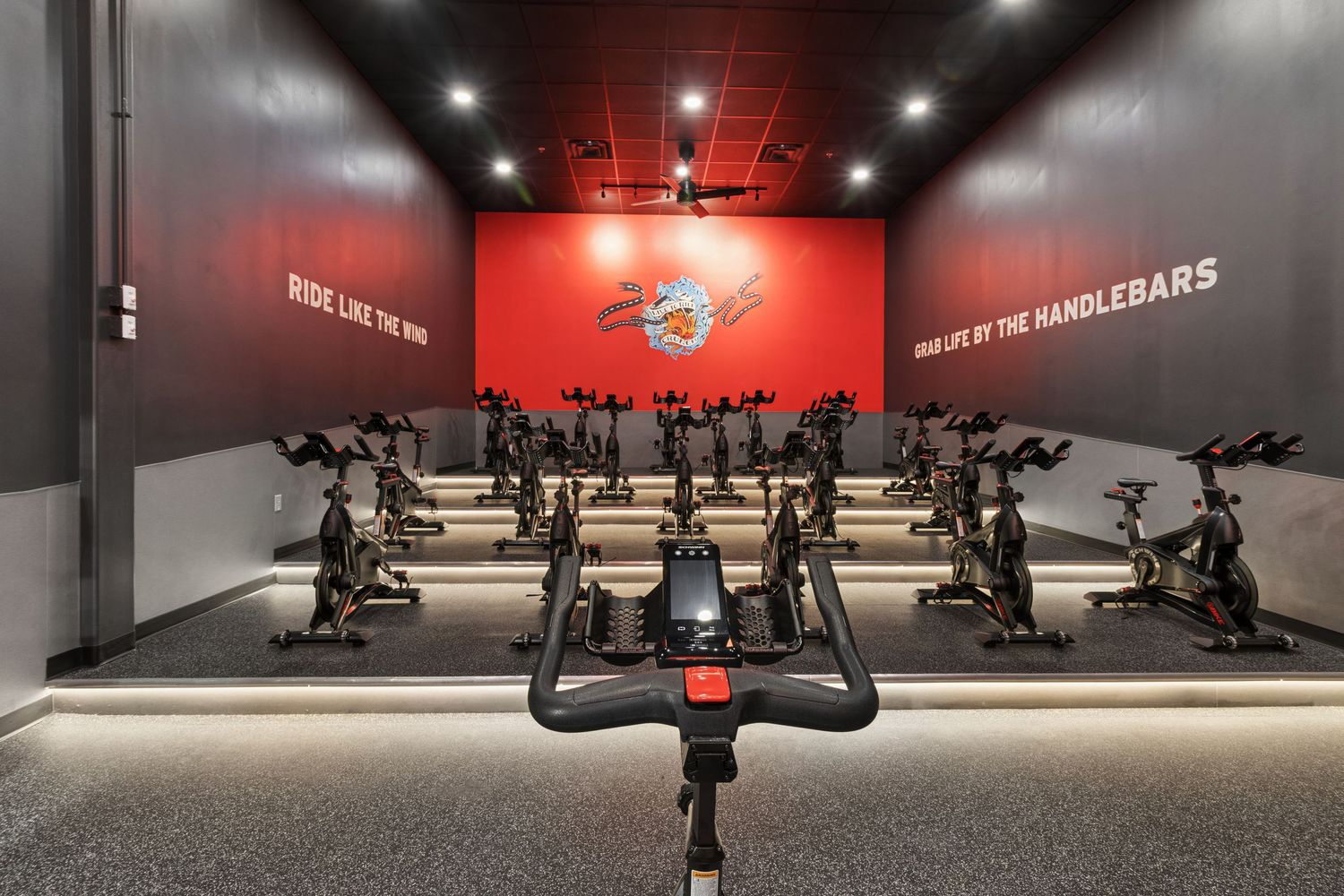 Indoor cycling studio with rows of stationary bikes, red accent wall, and motivational text in a modern gym setting.