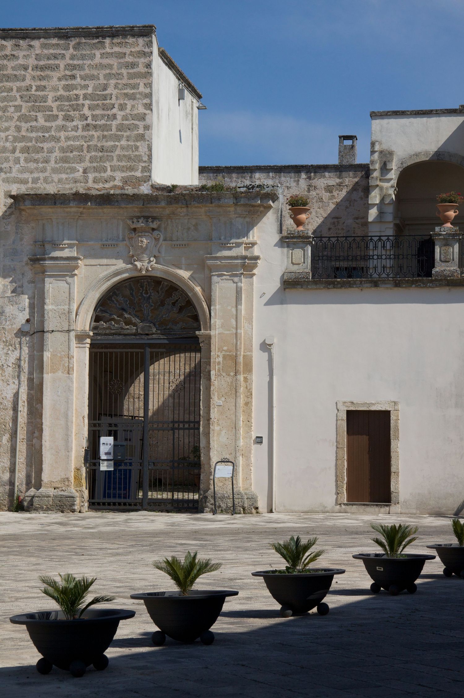 Historic stone courtyard with arched entrance gate, potted palm plants, and blue sky backdrop.