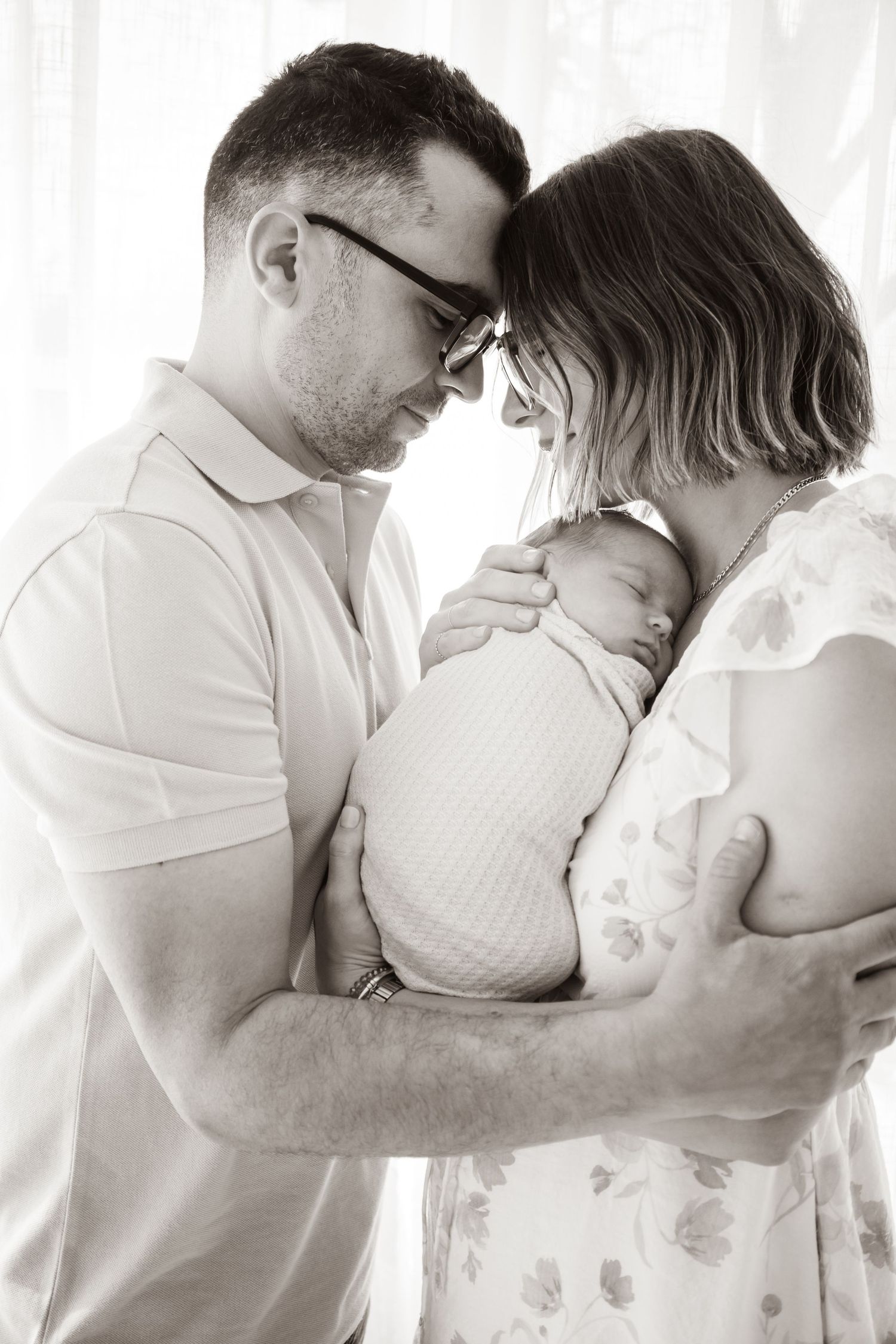 Black and white photo of parents tenderly holding their swaddled newborn baby, foreheads touching in an intimate moment.