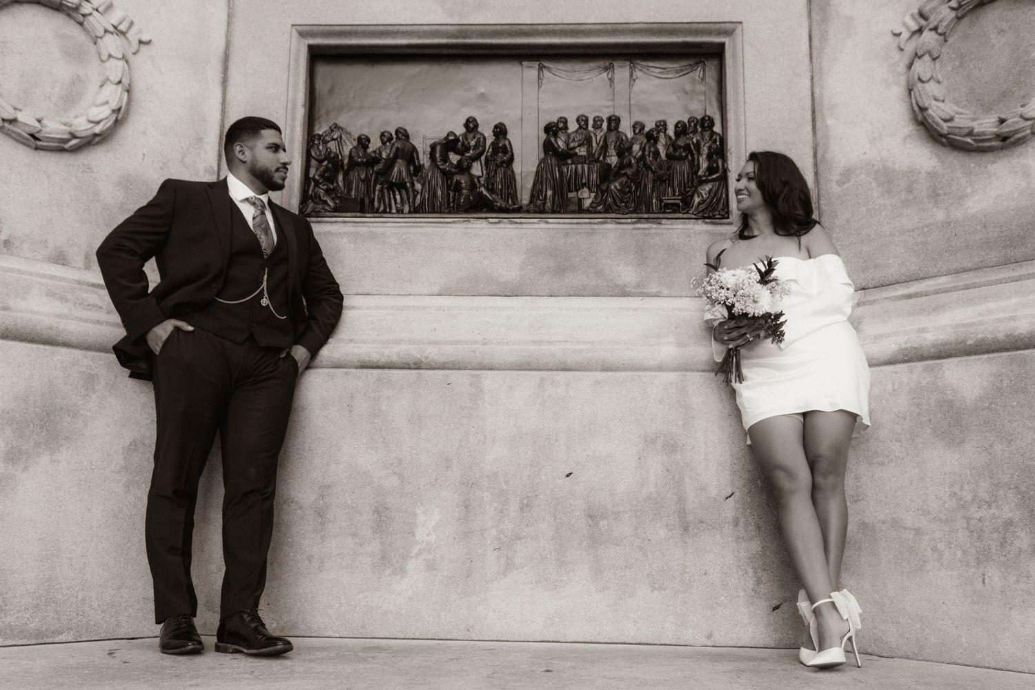 Black and white photo of a bride and groom posing apart against an ornate stone wall, exchanging glances.
