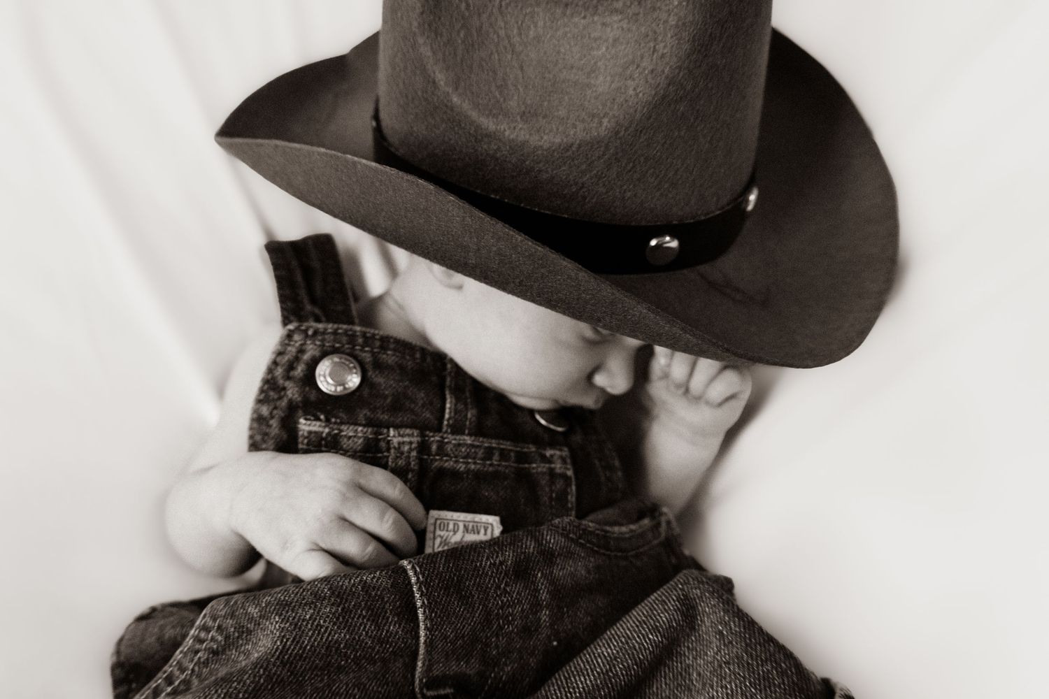 Adorable baby in overalls and oversized cowboy hat in black and white portrait photography.