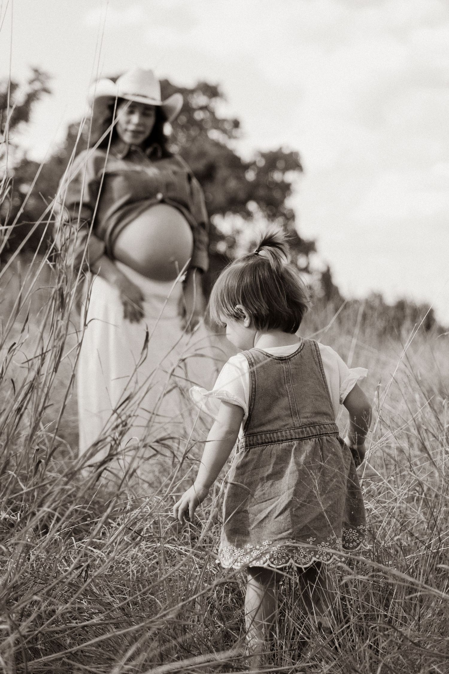 Black and white photo of a toddler walking toward her pregnant mother in a field of tall grass.