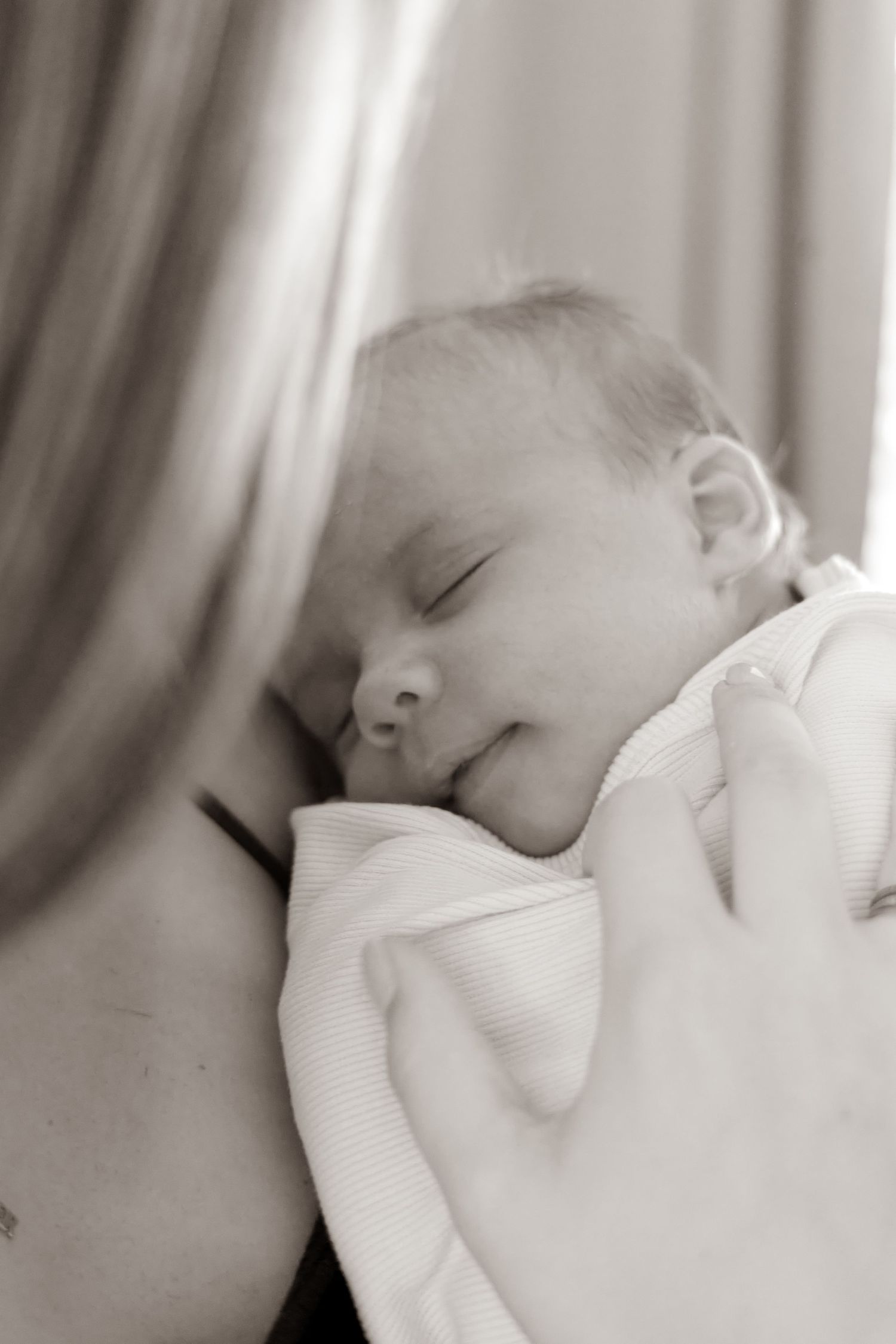 Black and white photo of a peaceful sleeping newborn baby cradled in a parent's arms, wrapped in a soft white blanket.