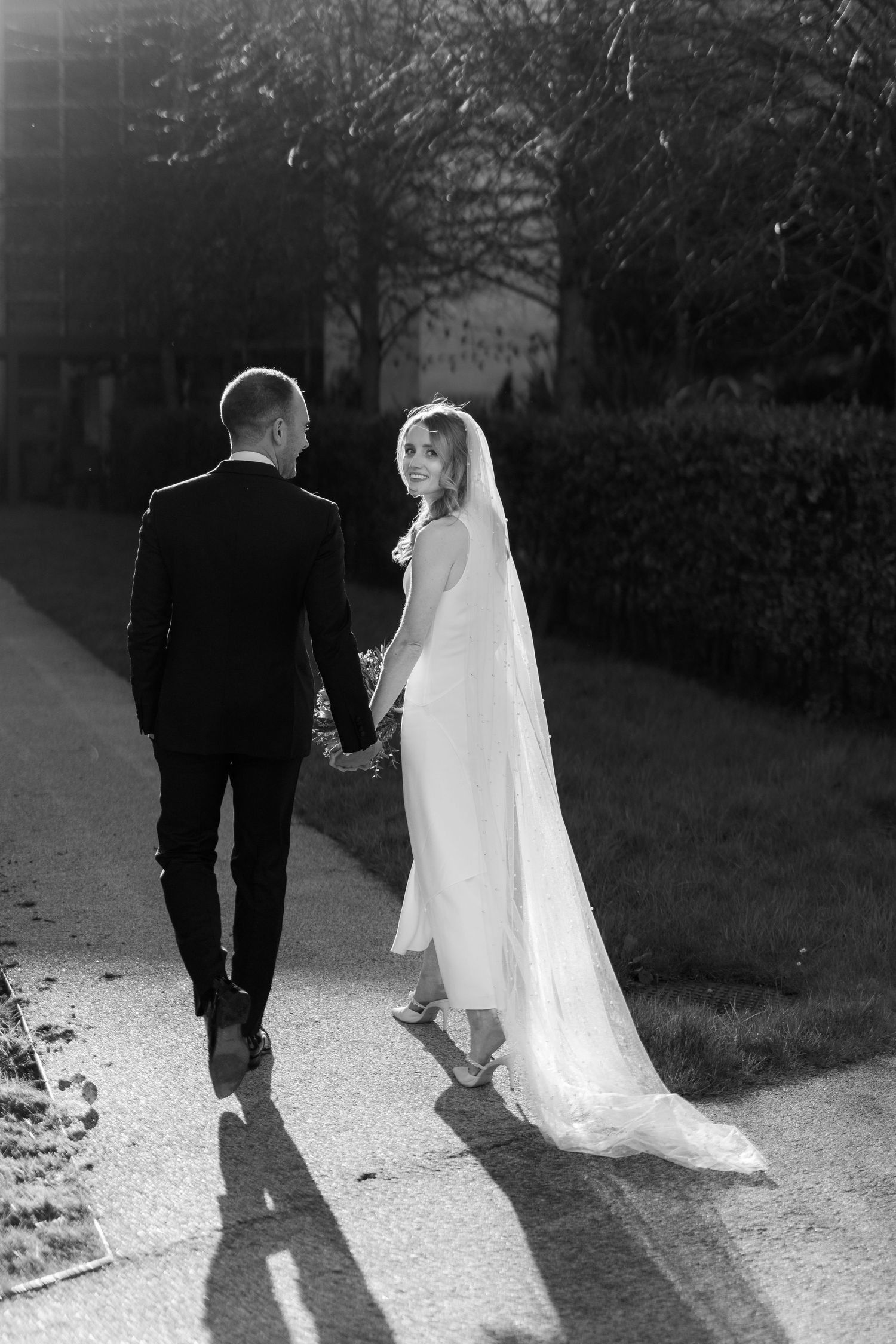 A couple walks hand in hand down a sunlit path with trailing wedding dress veil.