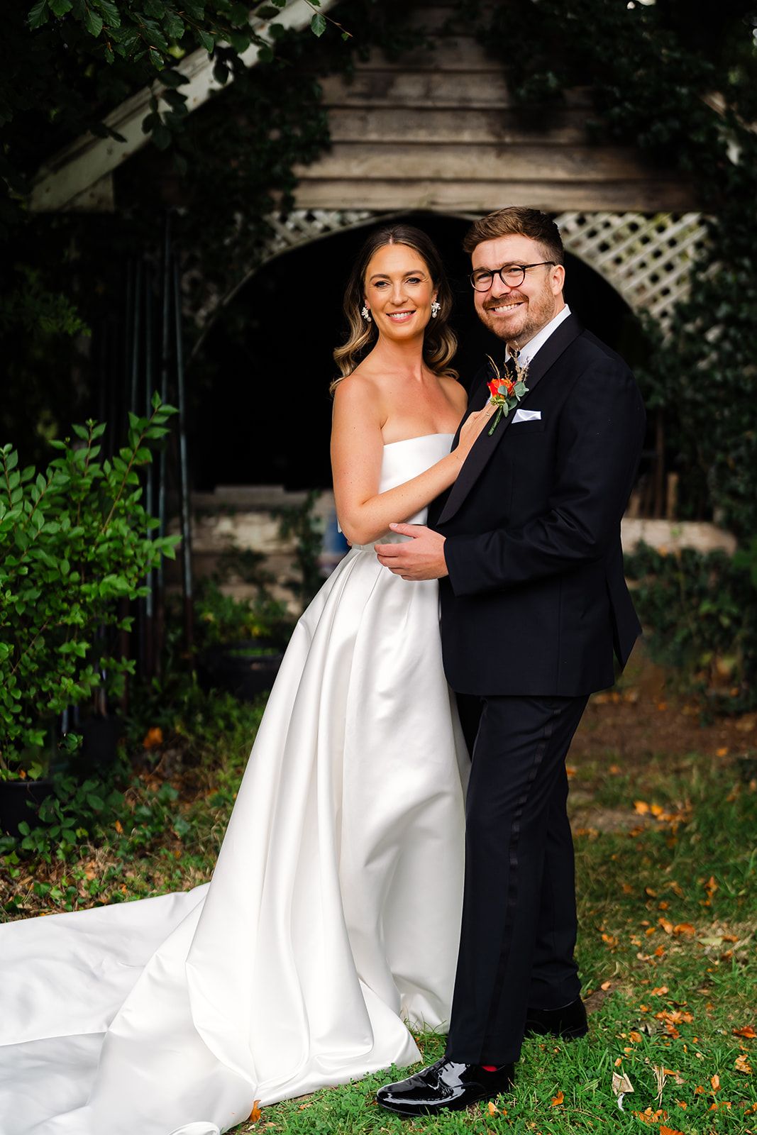A bride in a white strapless gown and groom in a black tuxedo embrace while standing in front of a garden arbor.