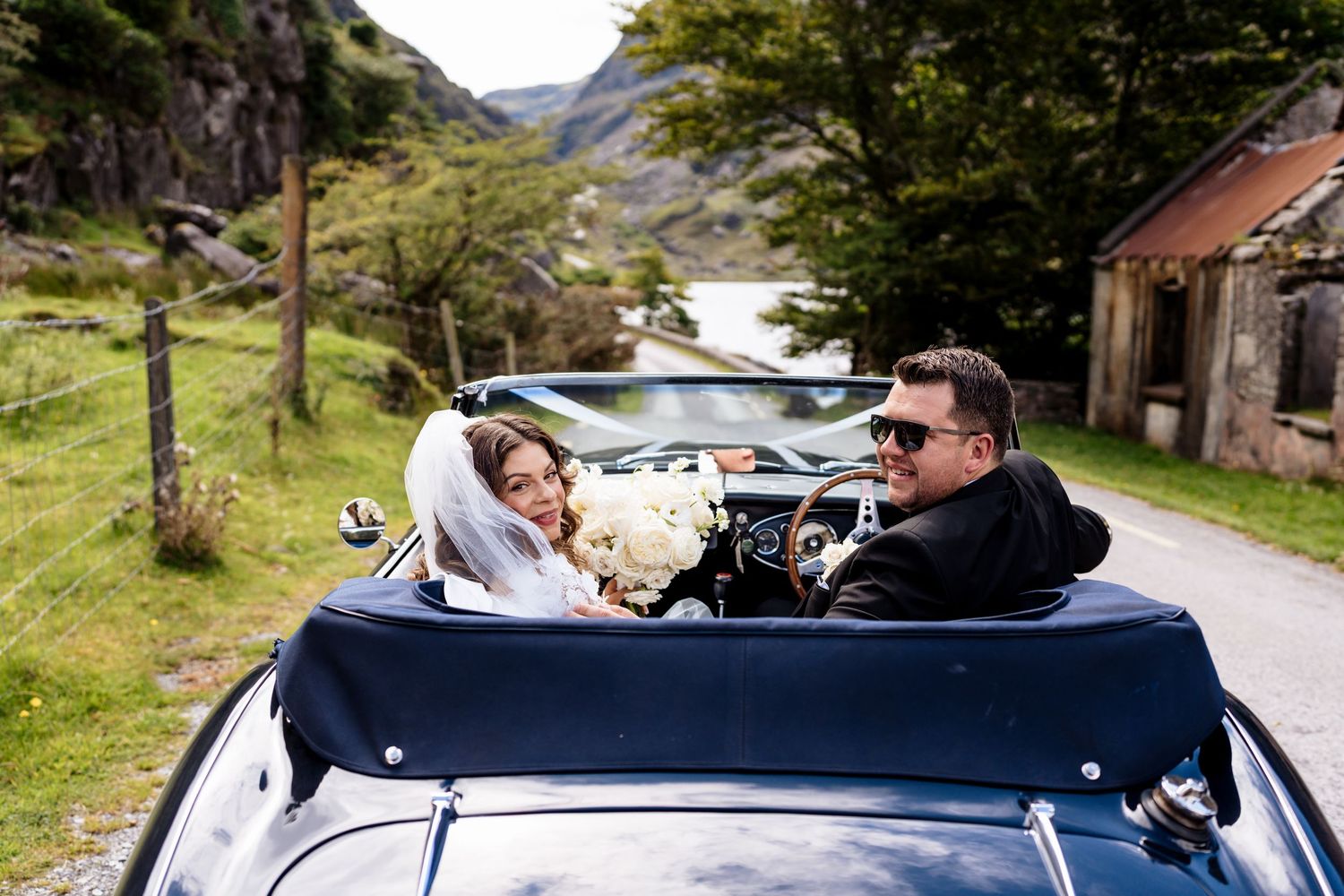 A group travels along a scenic mountain road in a convertible car on a sunny day.
