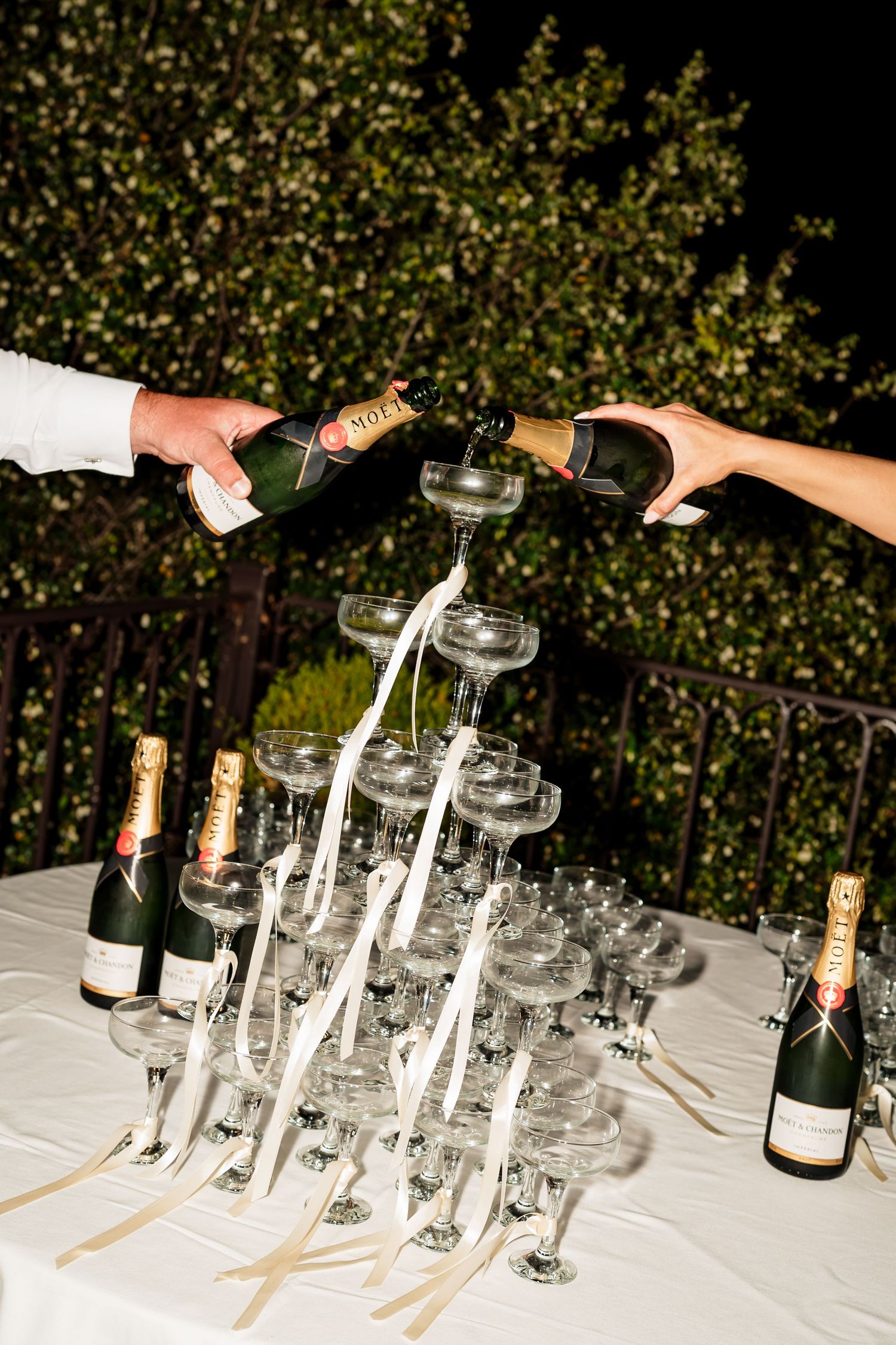 A sequence of champagne glasses being arranged in a tower formation on a white tablecloth outdoors.