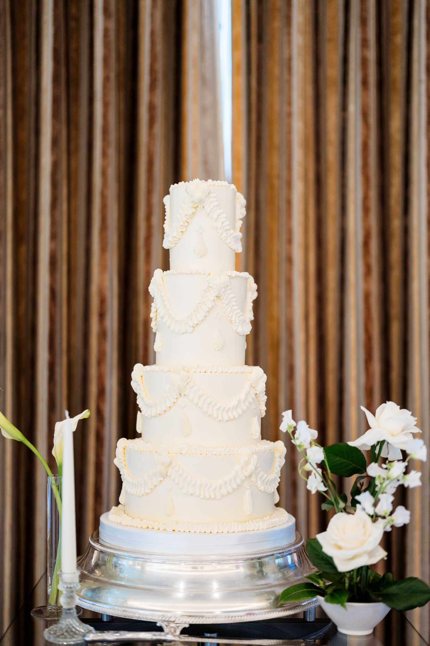 Three elegant white wedding cakes with delicate pearl details and floral decorations displayed on cake stands against wood panels.