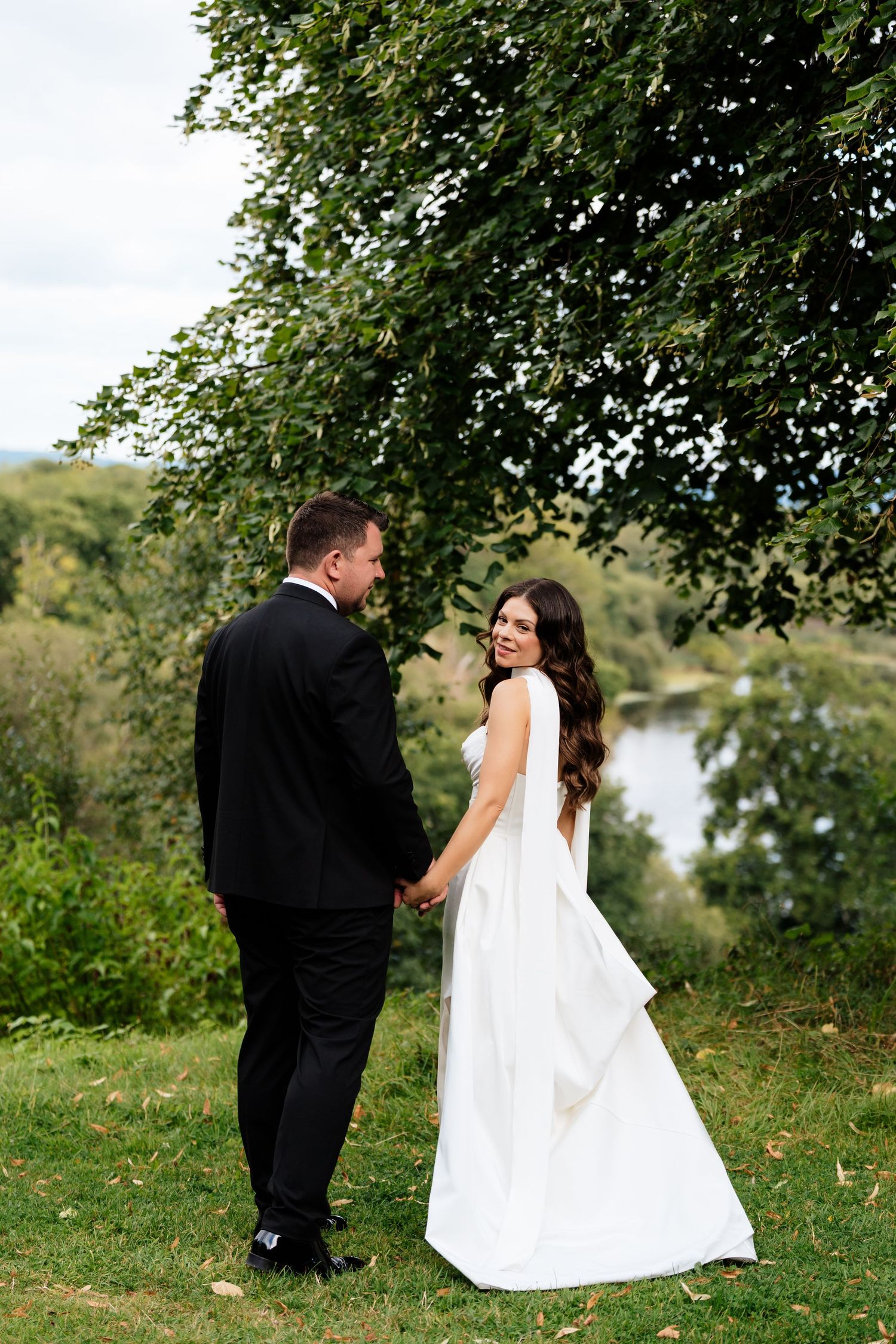 A couple in wedding attire stands on a grassy hill overlooking a lake under a tree canopy in a romantic outdoor setting.