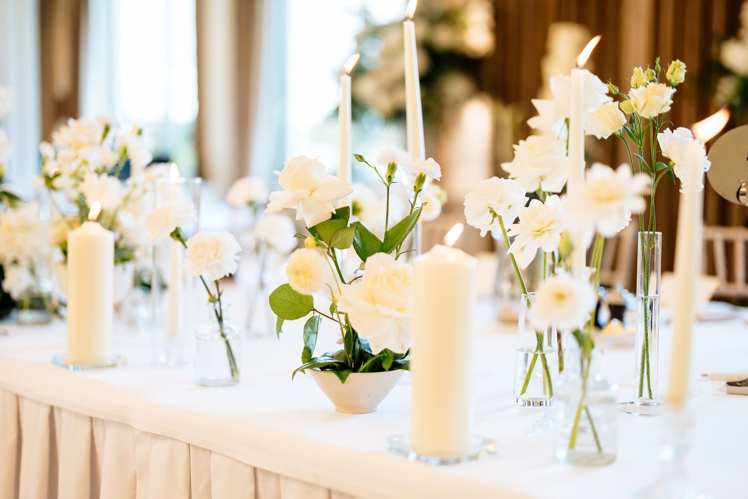 White floral centerpieces with candles line an elegant wedding reception table with soft glowing light.