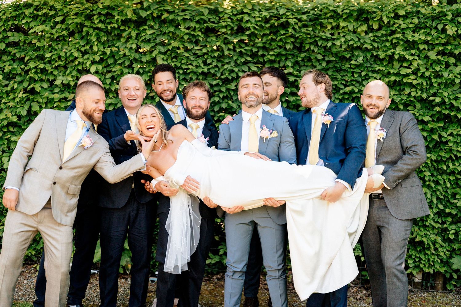 Wedding party poses playfully against an ivy wall with groomsmen holding a bride in a white dress.