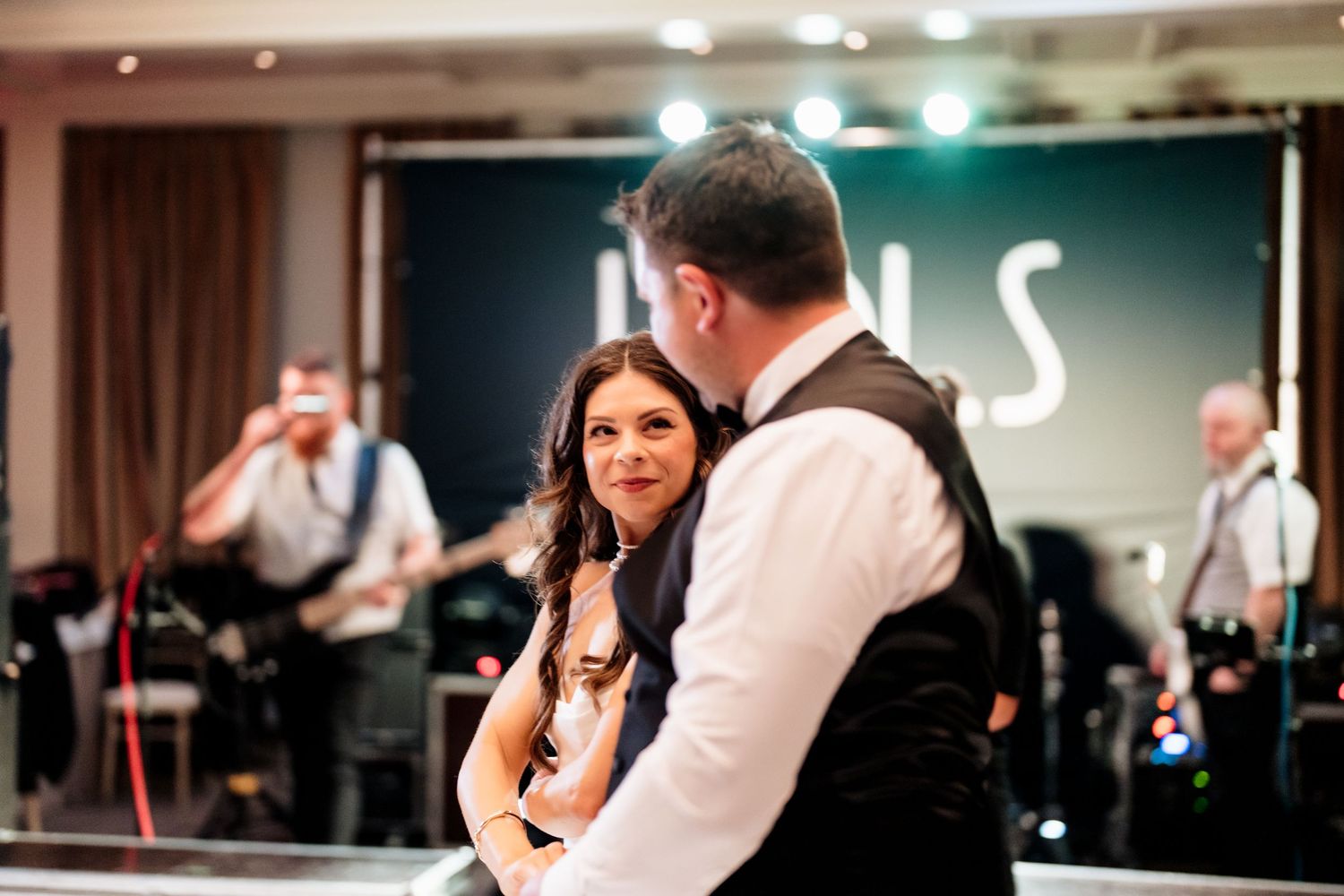 Wedding guests dance together to music at a lively indoor reception party with warm lighting and dark decor.