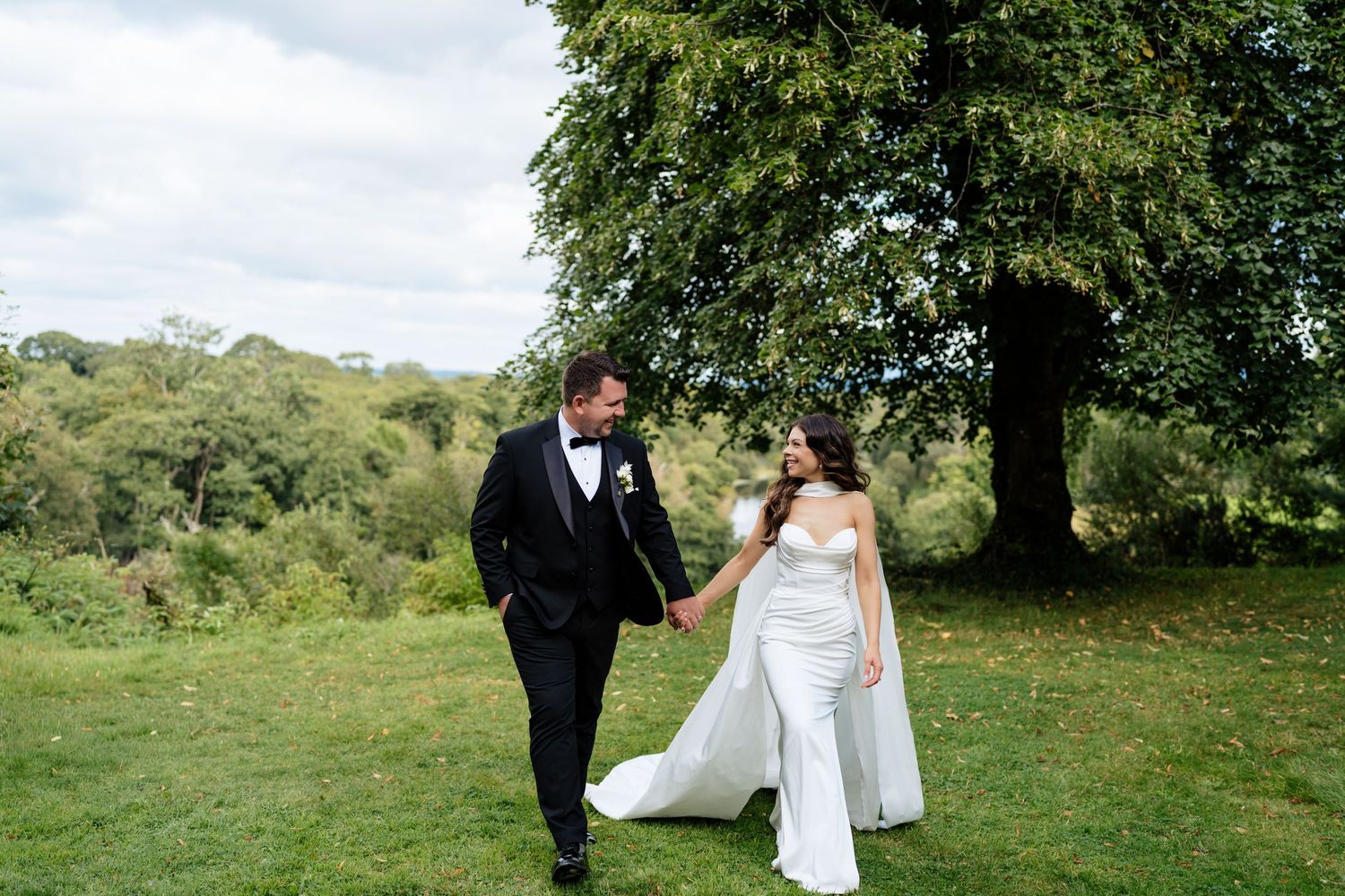 A romantic wedding couple walks together on lush green hills beneath cloudy skies on their special day.