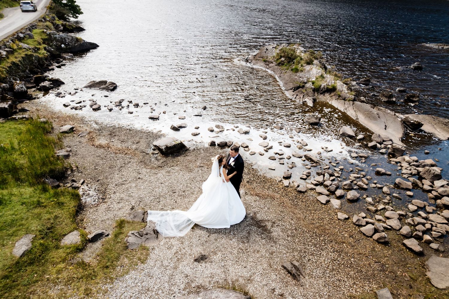 Aerial view of rocky coastline with couple on pebble beach overlooking blue water and evergreen trees.