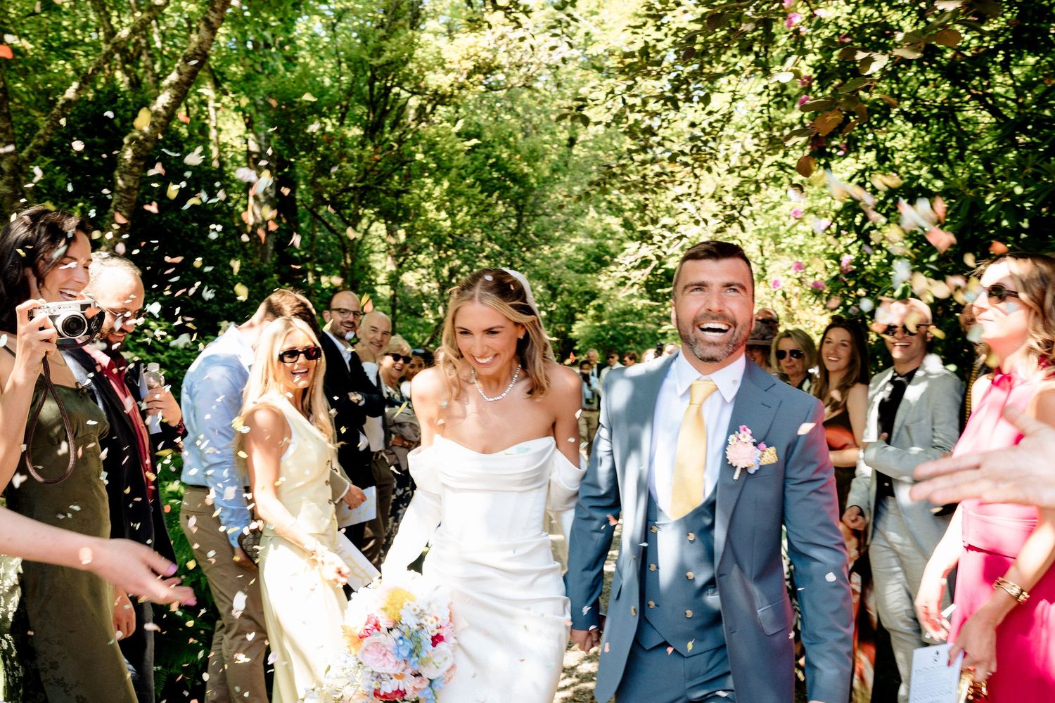 A joyful wedding celebration with guests throwing confetti over a couple walking down a garden path in summer sunlight.
