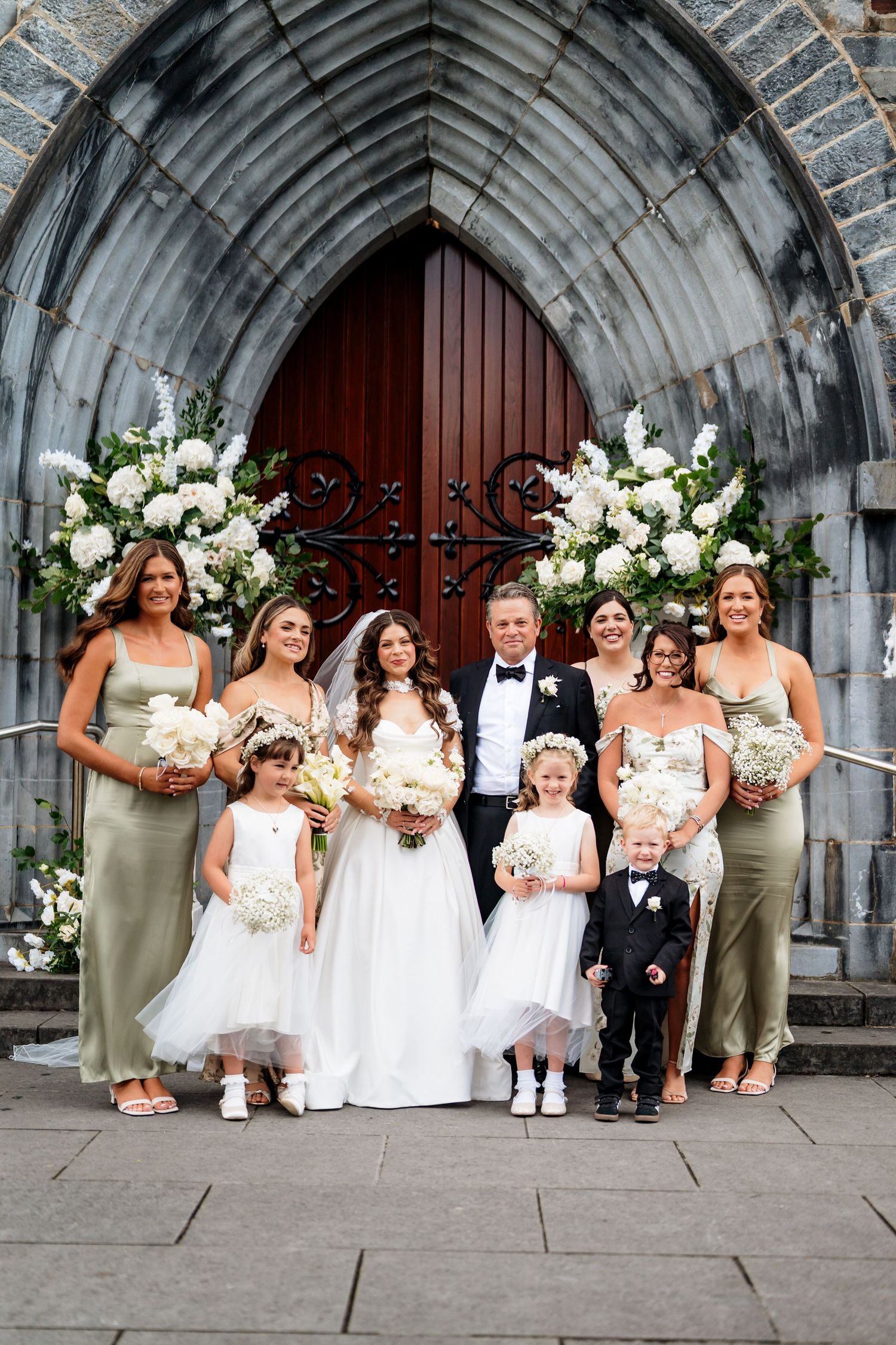 Wedding party in sage green and white dresses poses in front of a stone archway entrance.