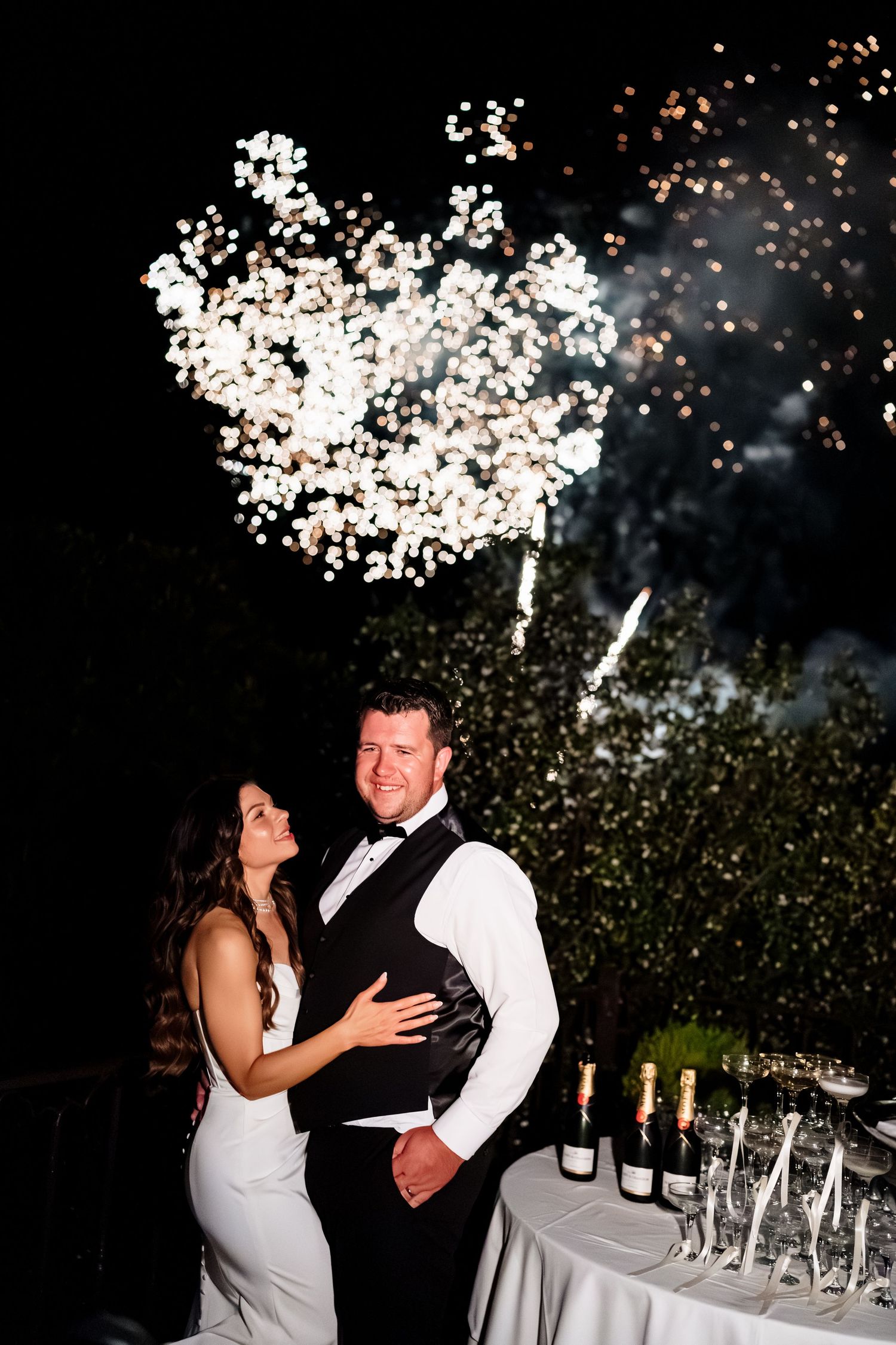A romantic newlywed couple shares their first dance outdoors at night with falling confetti under twinkling lights.