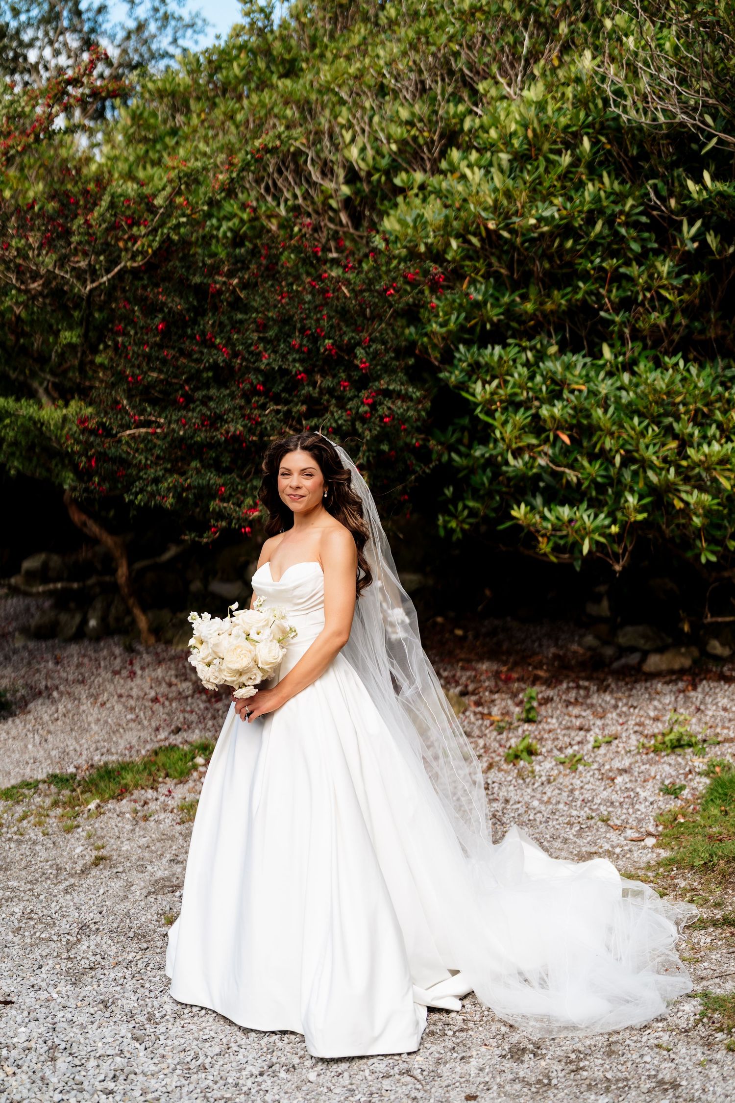 Bride in elegant white wedding gown poses gracefully on gravel path surrounded by lush greenery and flowering bushes.