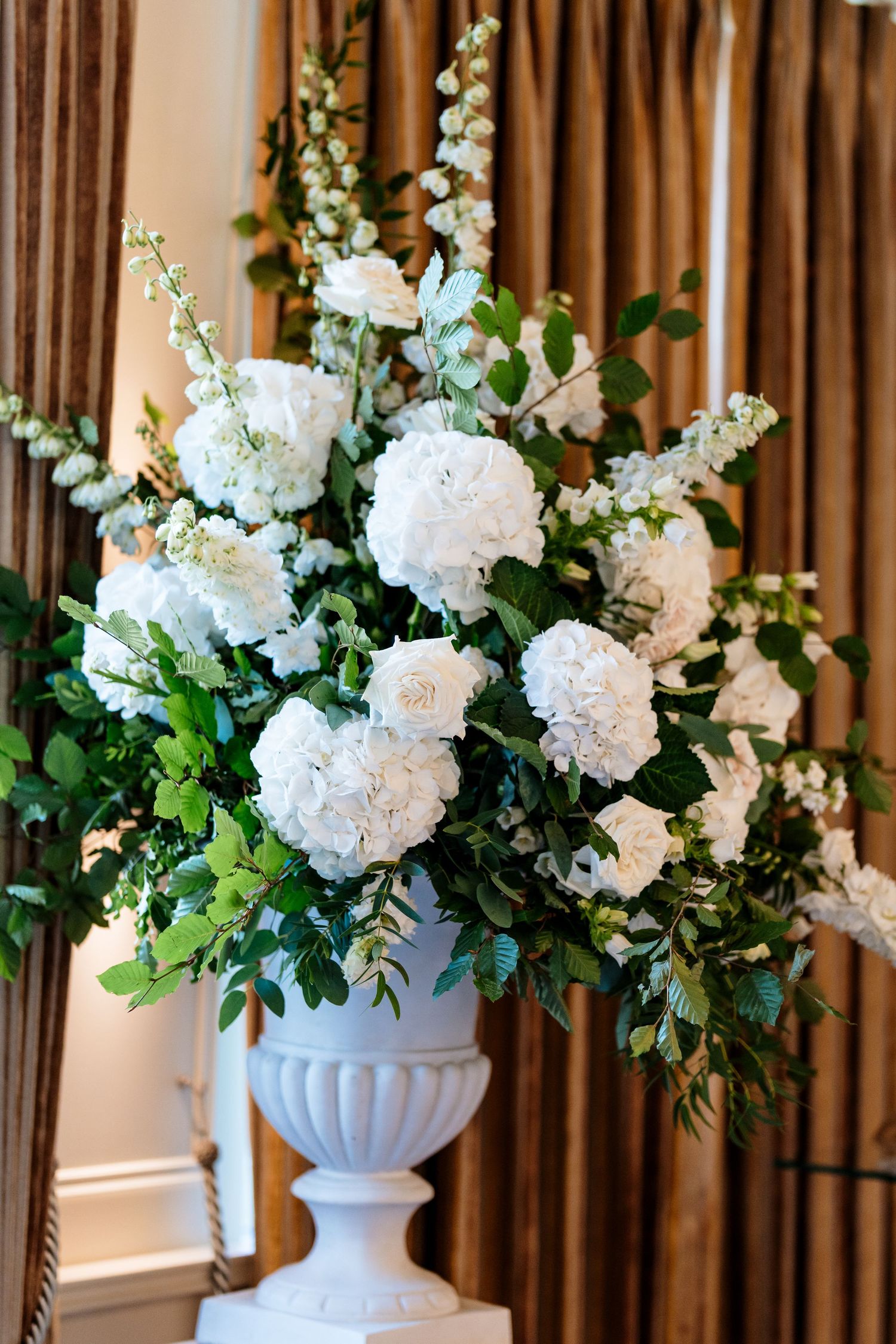 Elegant white floral arrangements in classic urns with fresh greenery decorate a formal indoor space.