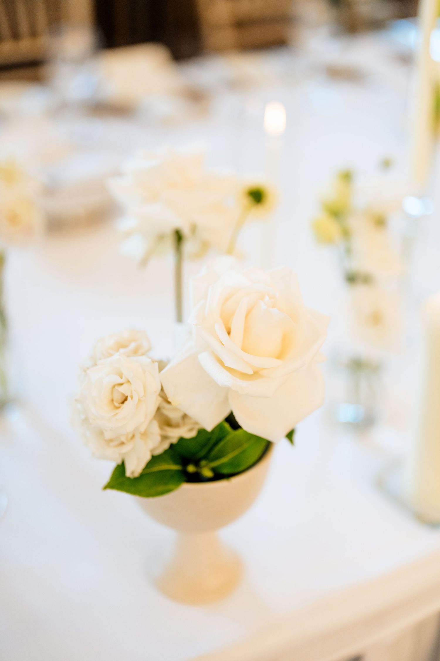 Elegant wedding table centerpiece with delicate white and blush roses in a minimalist vase against a soft blurred background.