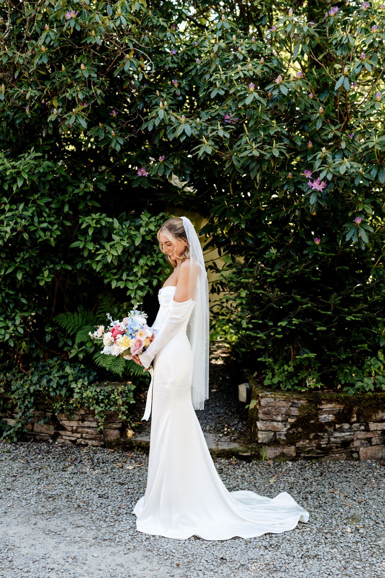 A bride in a long white gown and veil stands against a lush garden backdrop while holding a colorful floral bouquet.