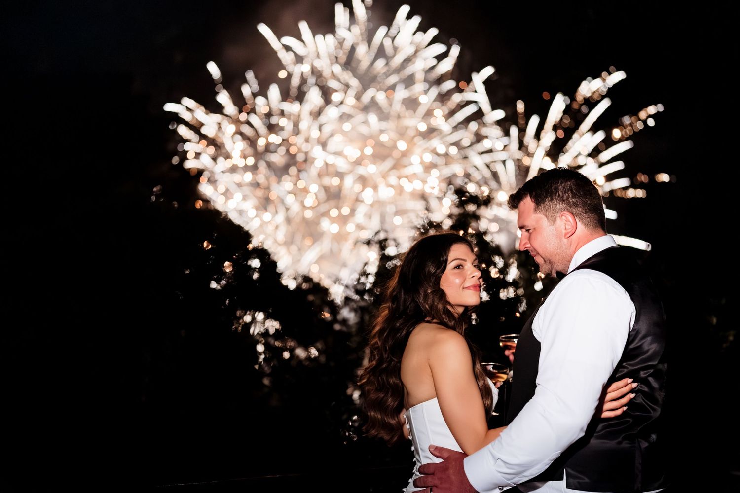 A couple shares a romantic moment under bursting fireworks at their wedding celebration during nighttime.