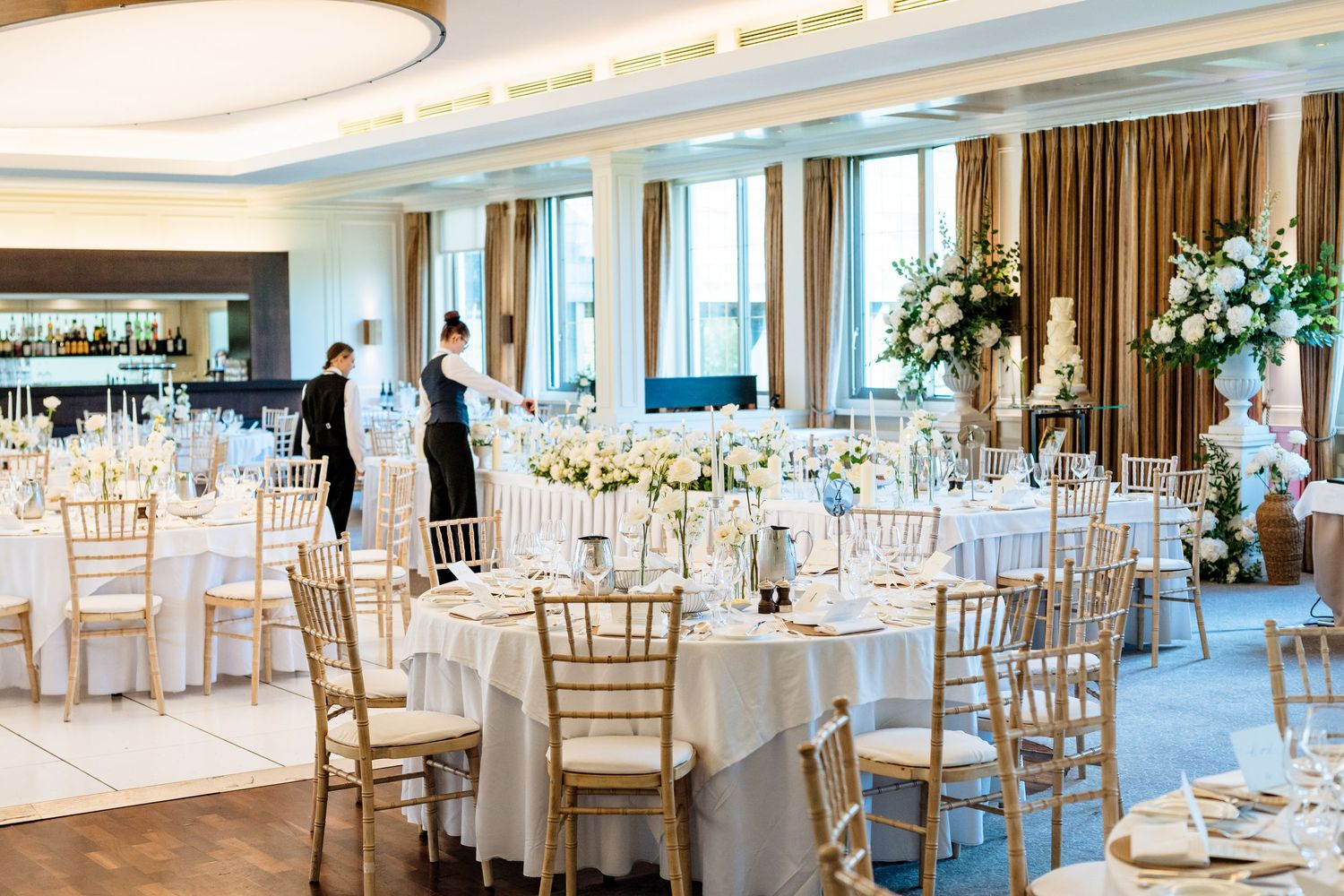 Elegant wedding reception venue with white tablecloths, gold chairs, and large windows overlooking water views.