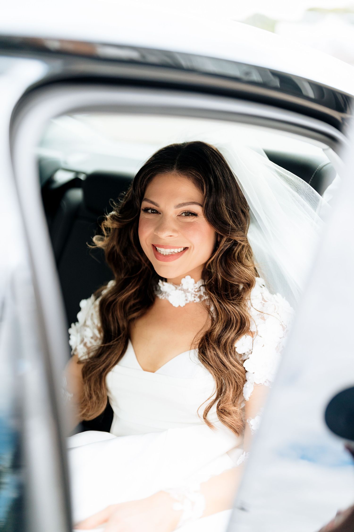 Portrait of a smiling woman with wavy brown hair wearing white in her car with natural backlighting.