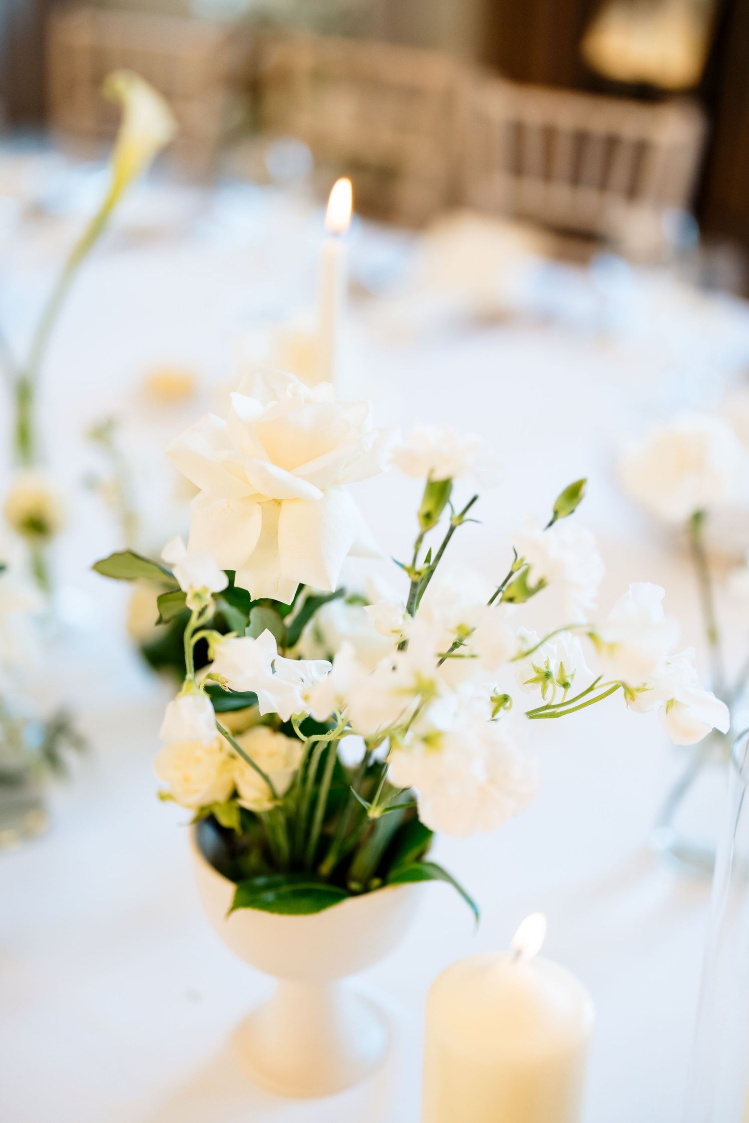 Elegant white floral centerpiece with roses and delicate blooms in a white vase on a crisp tablecloth with soft candlelight.