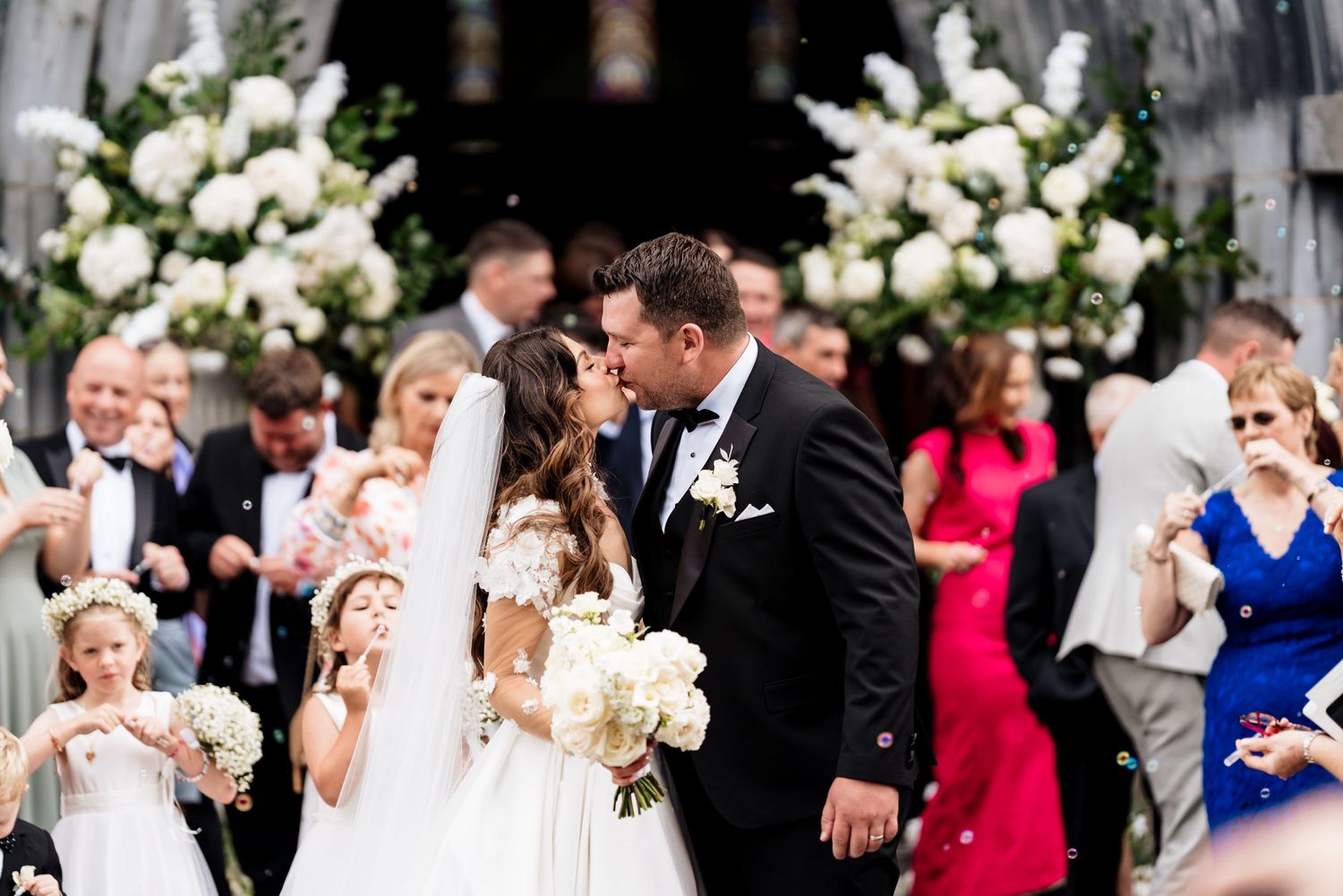 Wedding party celebrates during joyful outdoor ceremony with white floral arrangements and formal attire.