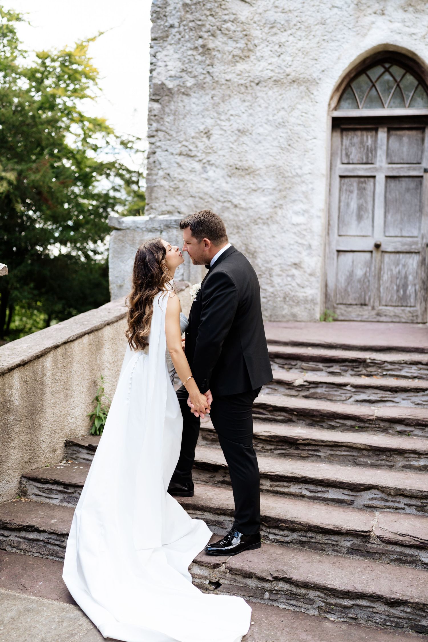 A romantic couple in wedding attire share an intimate moment on stone steps outside a historic building with arched windows.