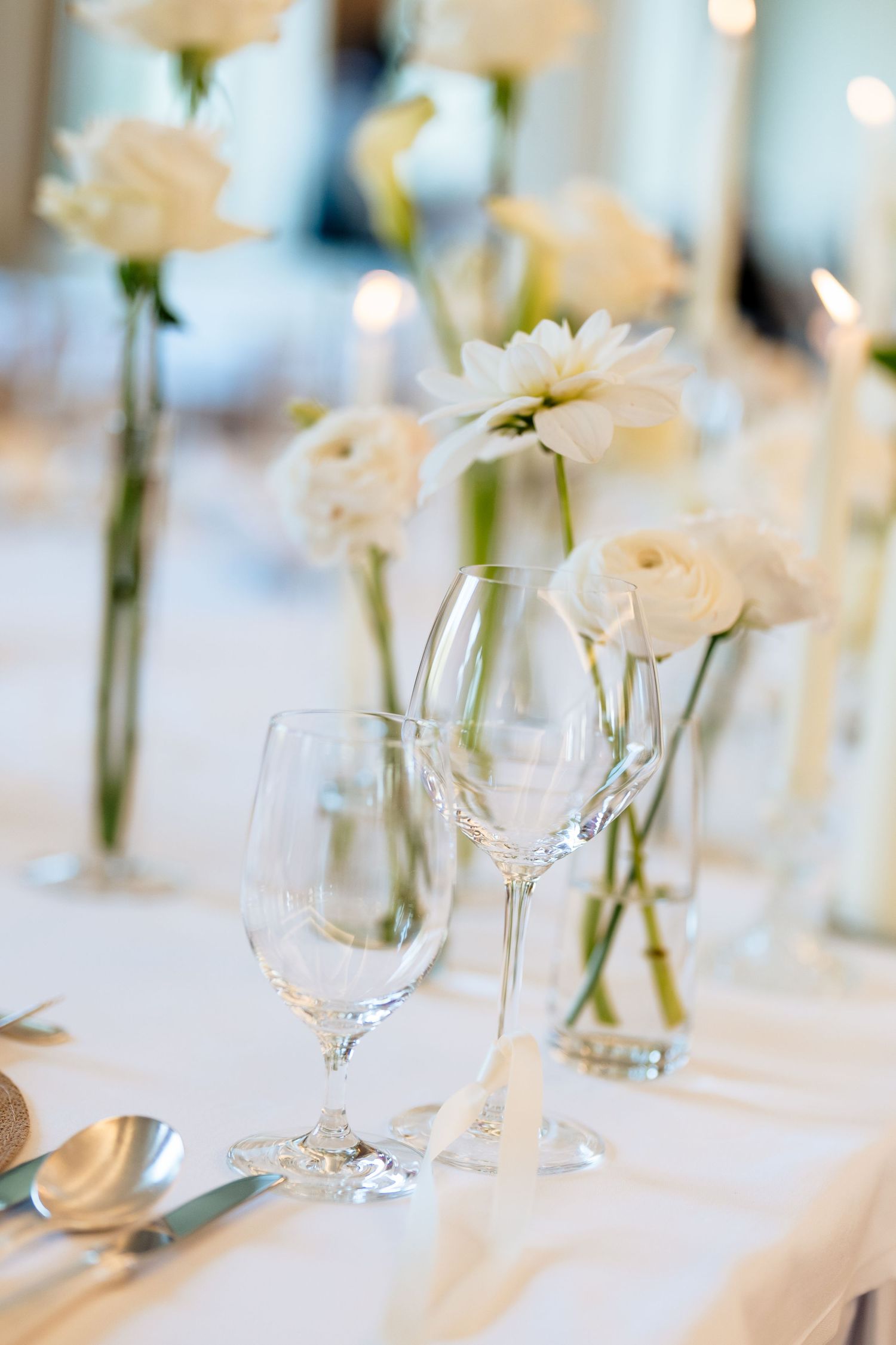 Elegant table setting with white roses in bud vases and crystal wine glasses on crisp white tablecloth.