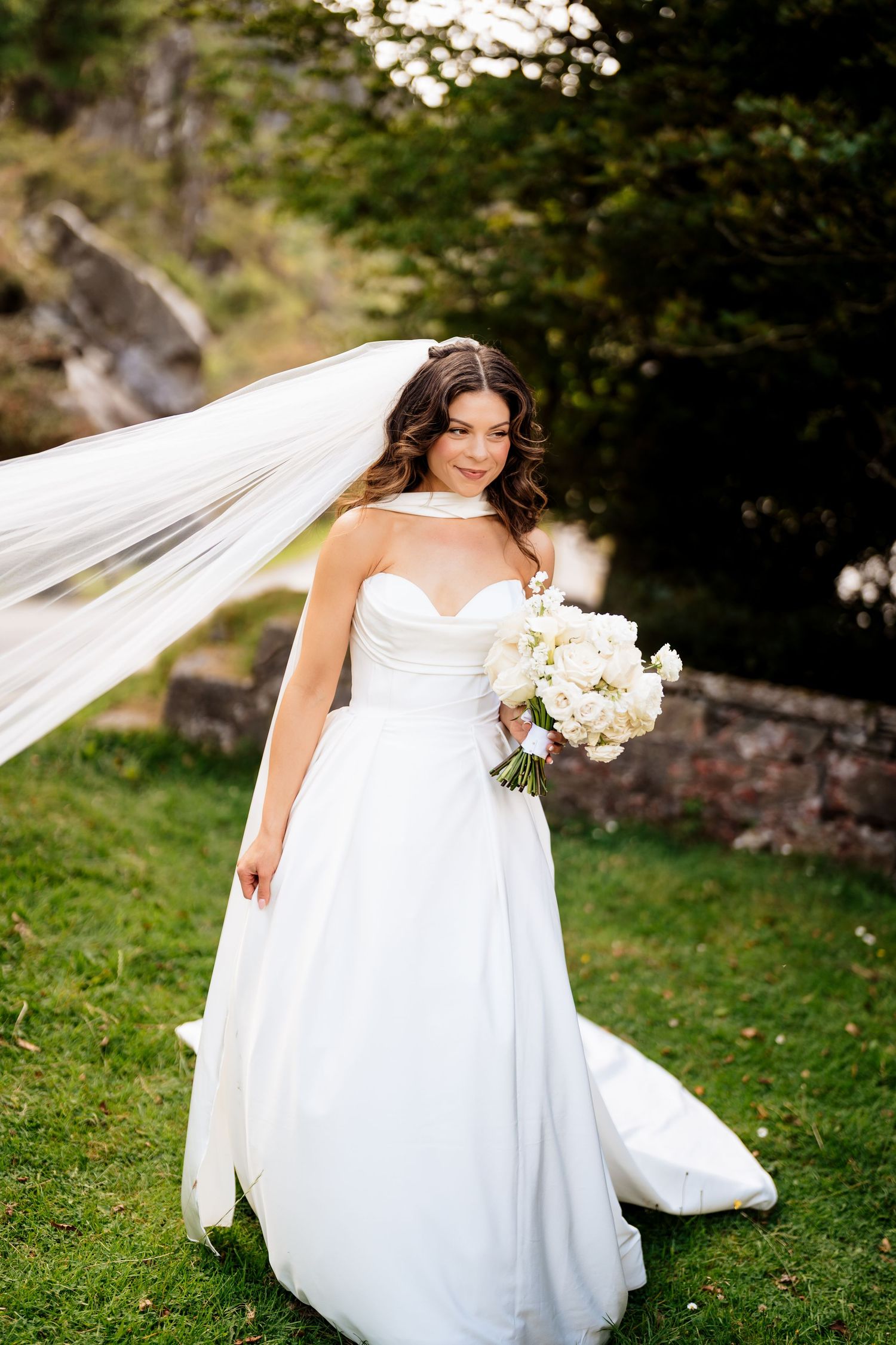 Elegant white wedding gown with sweetheart neckline and flowing veil captured outdoors with white bouquet and natural scenery.