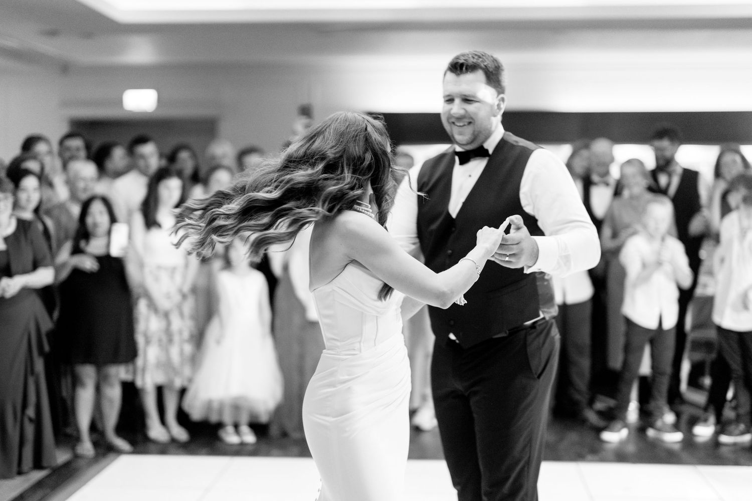 A couple dances together at their wedding reception as guests look on in a black and white photograph.