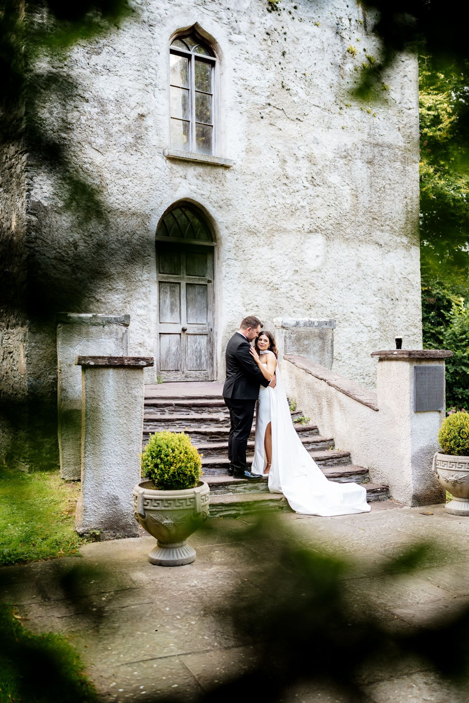 Romantic couple embracing on stone steps in front of a rustic white building with dark shadows framing the scene.