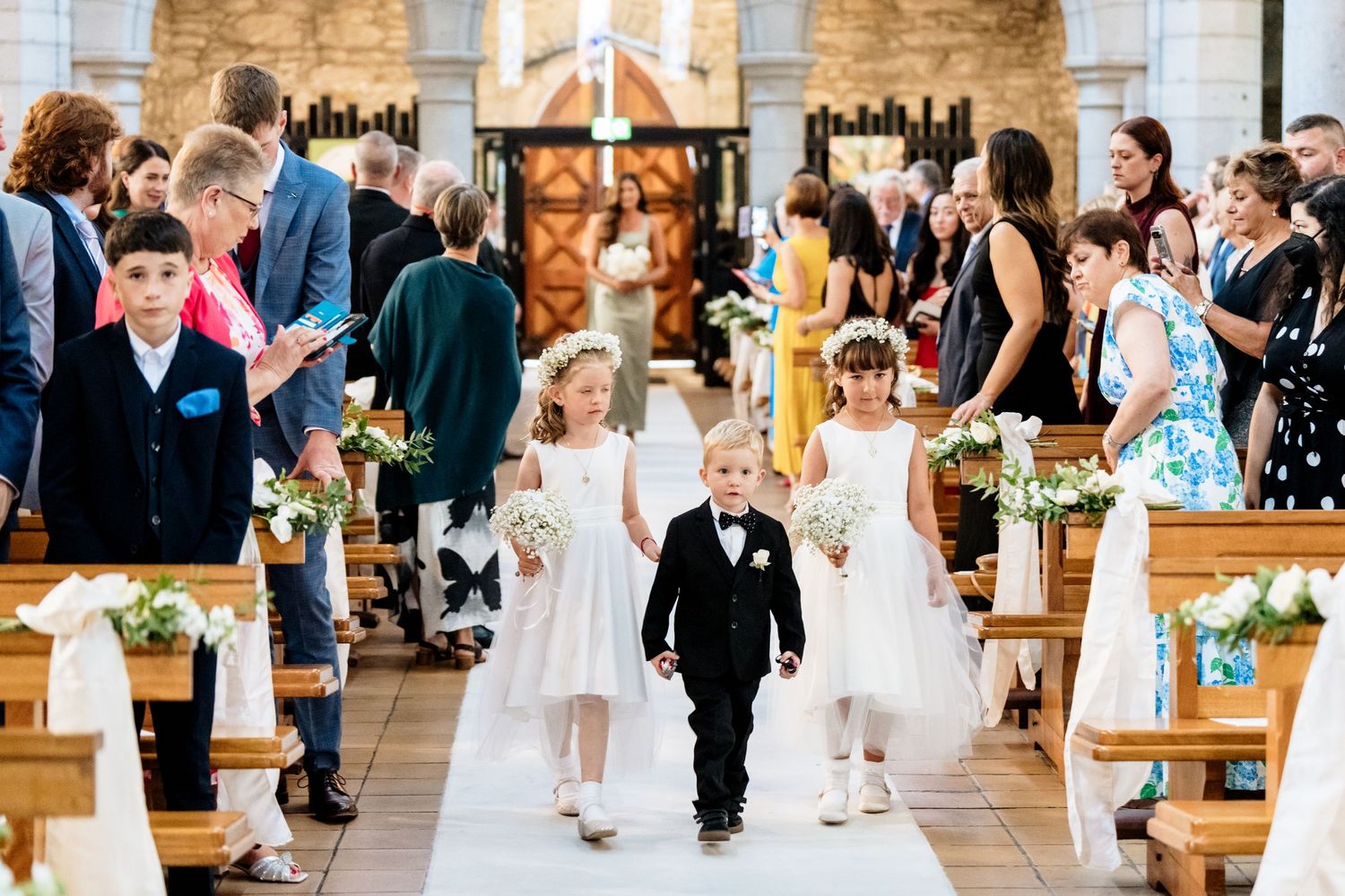 Wedding party of children in formal white dresses and dark suits walk down aisle in church during ceremony.