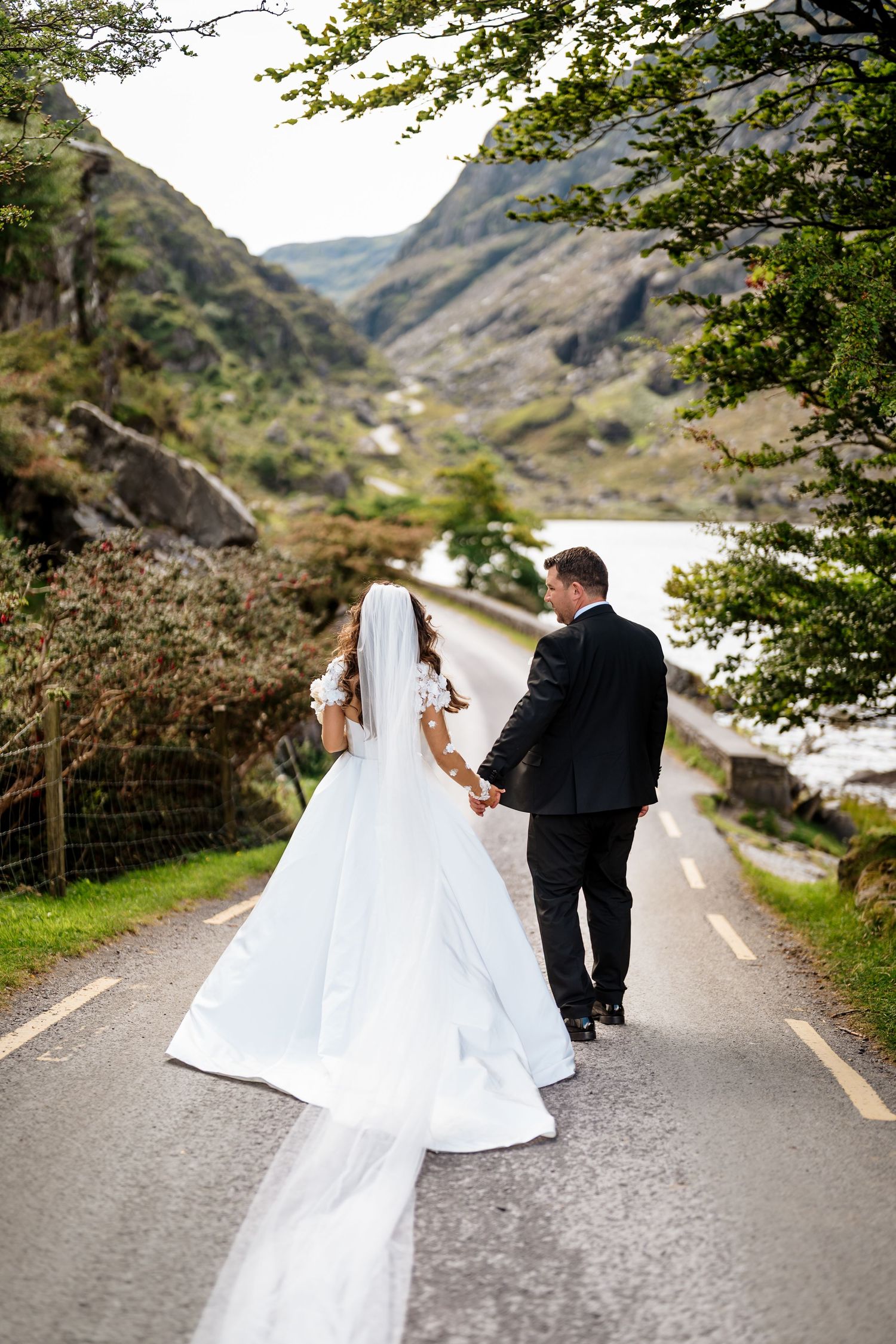 Newlywed couple walks hand in hand on scenic mountain road framed by lush greenery and dramatic cliffs.
