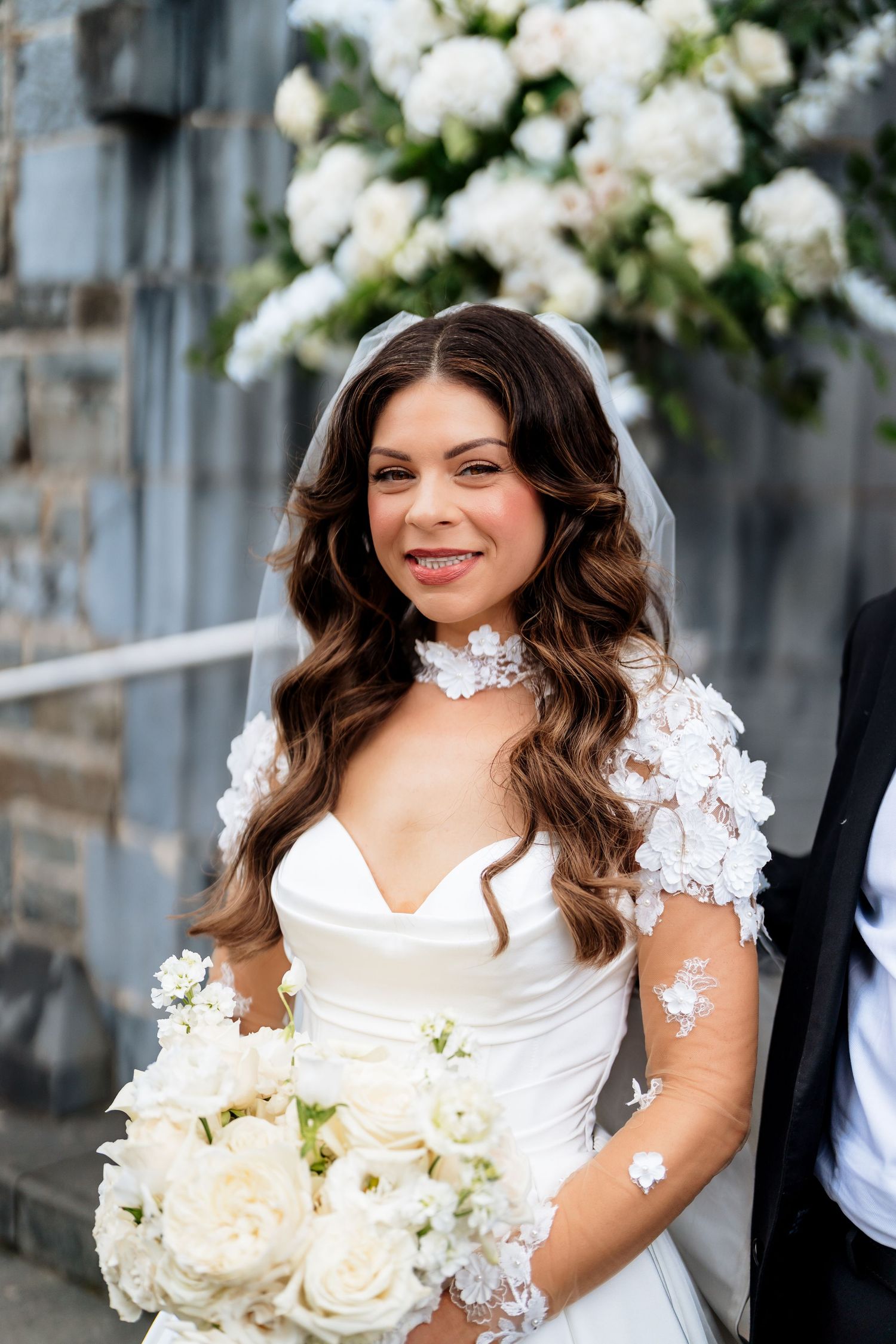 A joyful bride in a white lace dress holds a bouquet of cream roses against a blurred outdoor background.