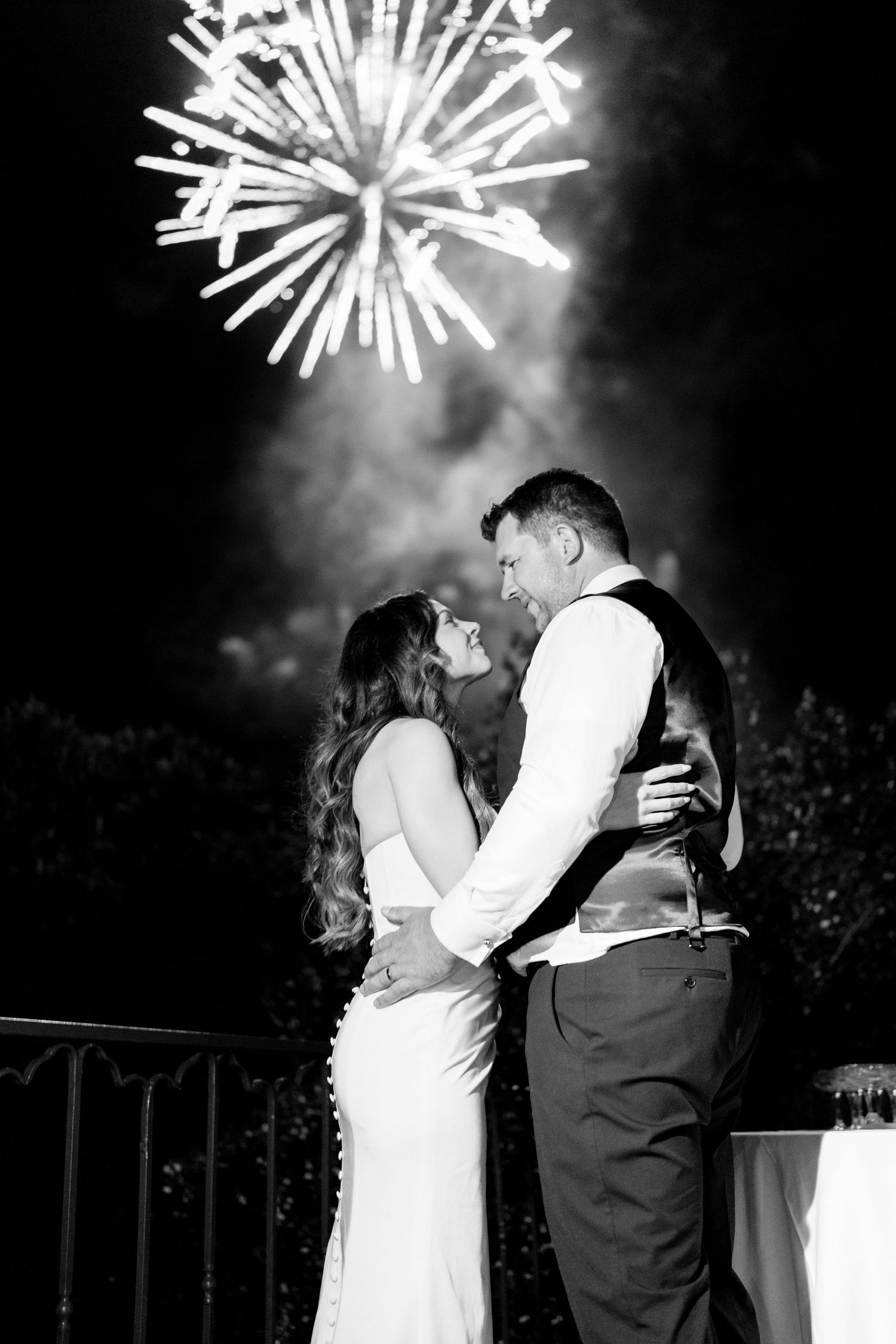 A couple shares a romantic kiss under bursting fireworks during a nighttime celebration with a dramatic black and white effect.